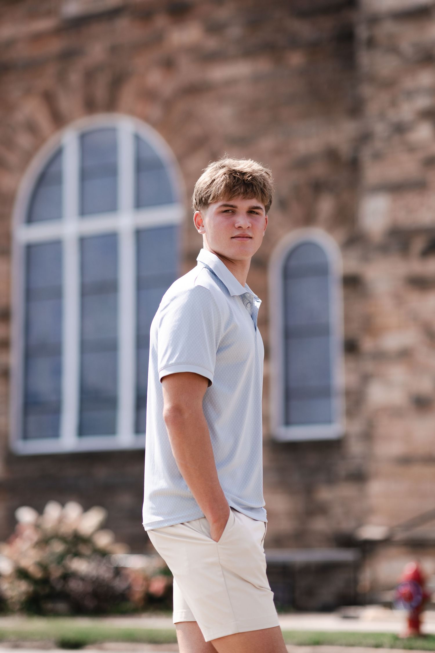 Young man in light blue polo and white shorts standing outdoors near a stone building with arched windows.