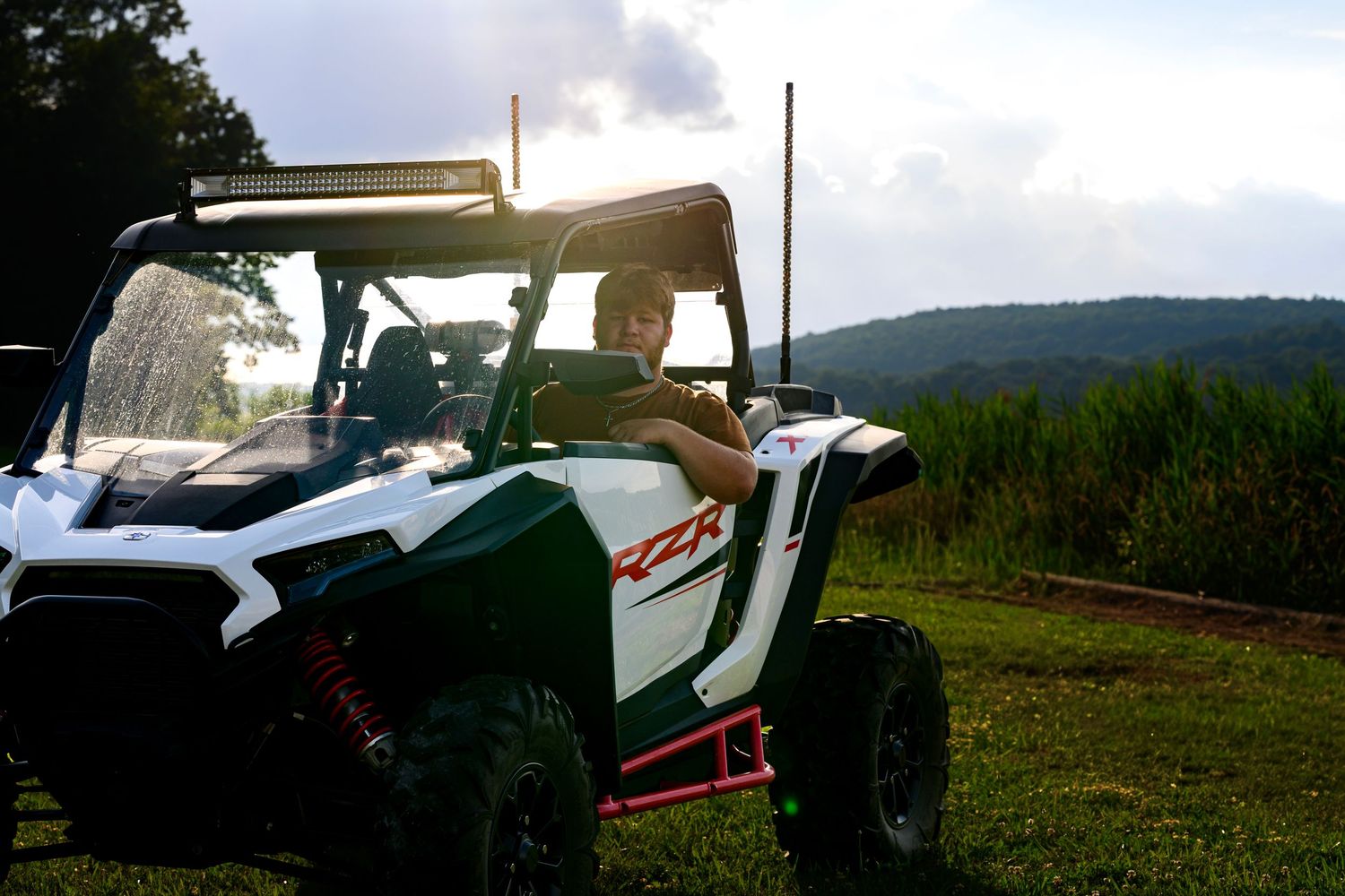 Person driving a white and green Polaris RZR off-road UTV on a grassy trail with scenic hills in background.