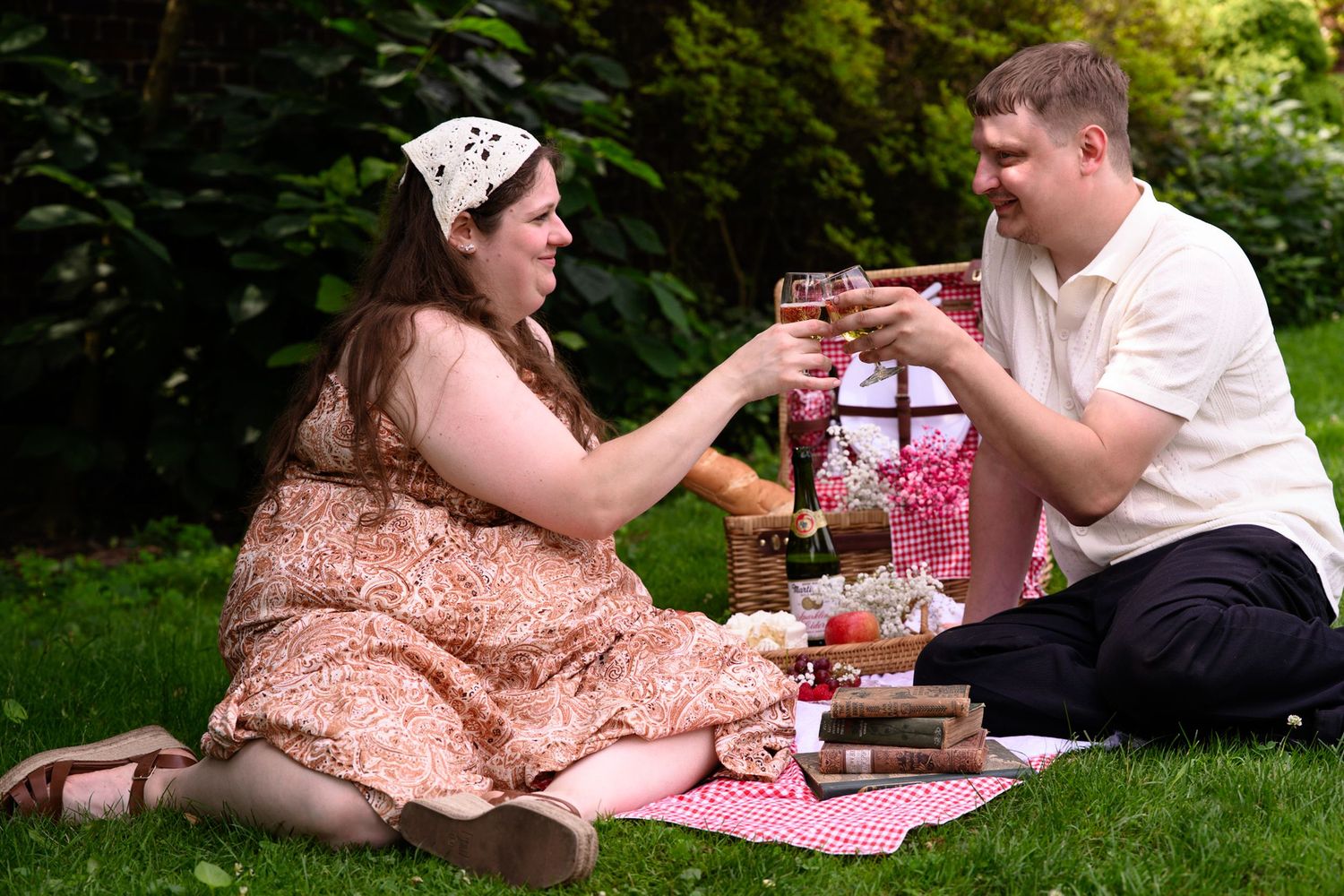 Couple enjoying a romantic outdoor picnic on a blanket, toasting wine glasses surrounded by books and a gift basket.