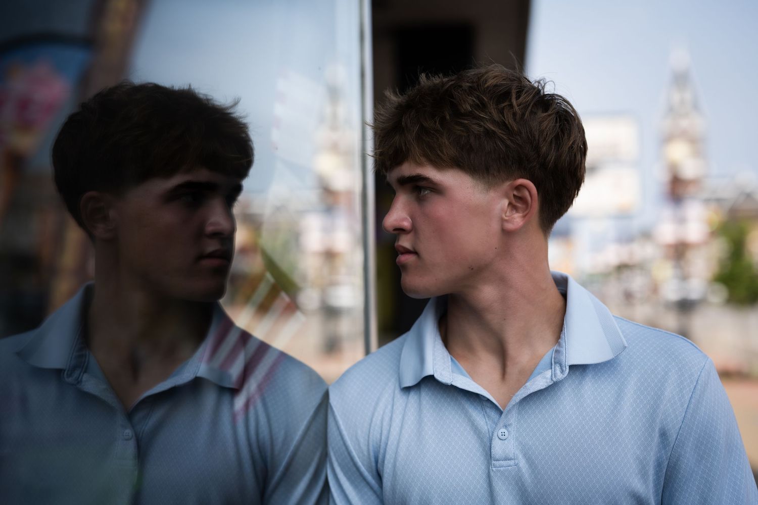 Young man in blue polo shirt gazes sideways, his reflection visible in a glass window outdoors.