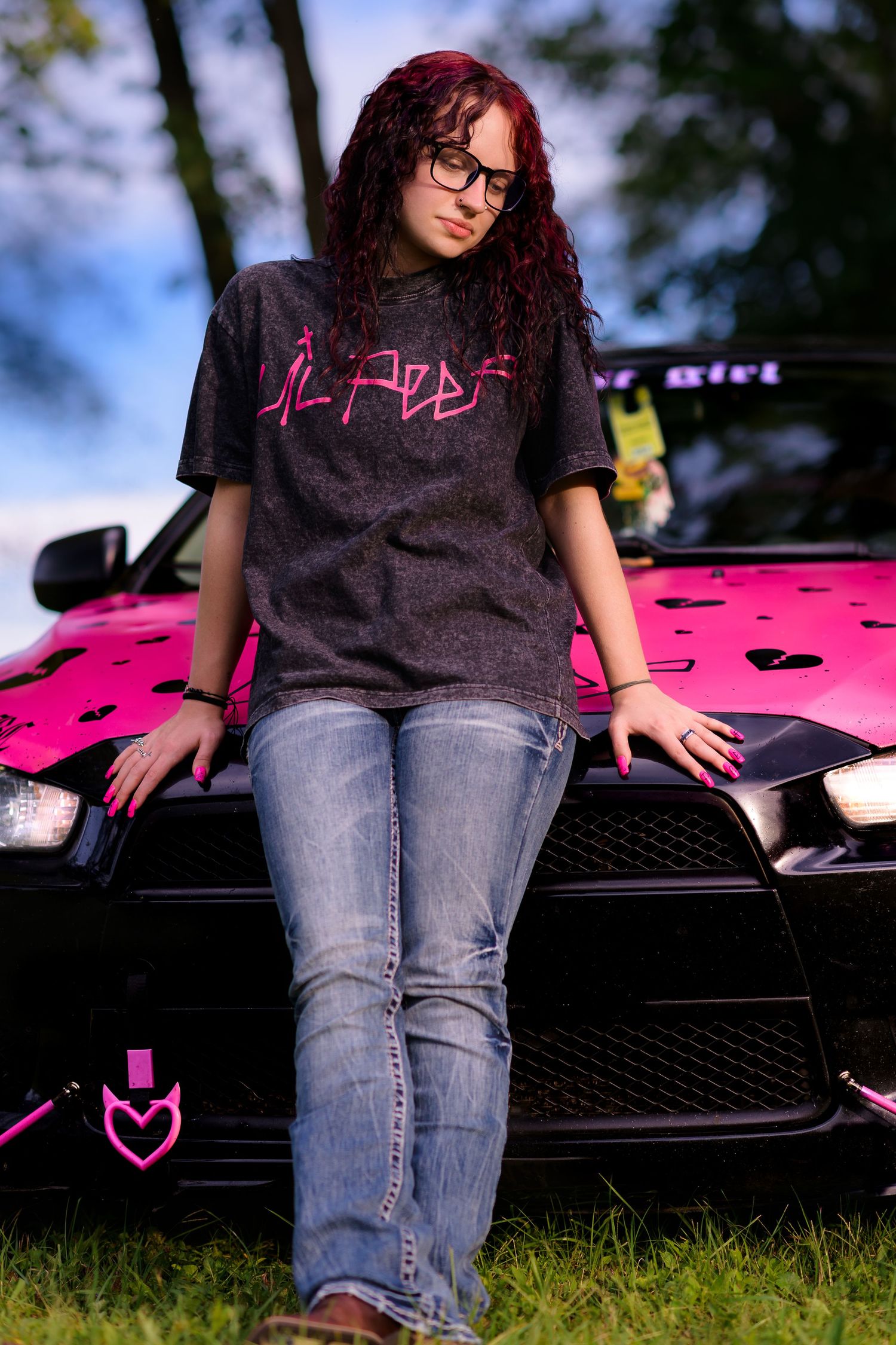 Young woman with red hair and glasses leaning on a pink and black car wearing a graphic tee and jeans.