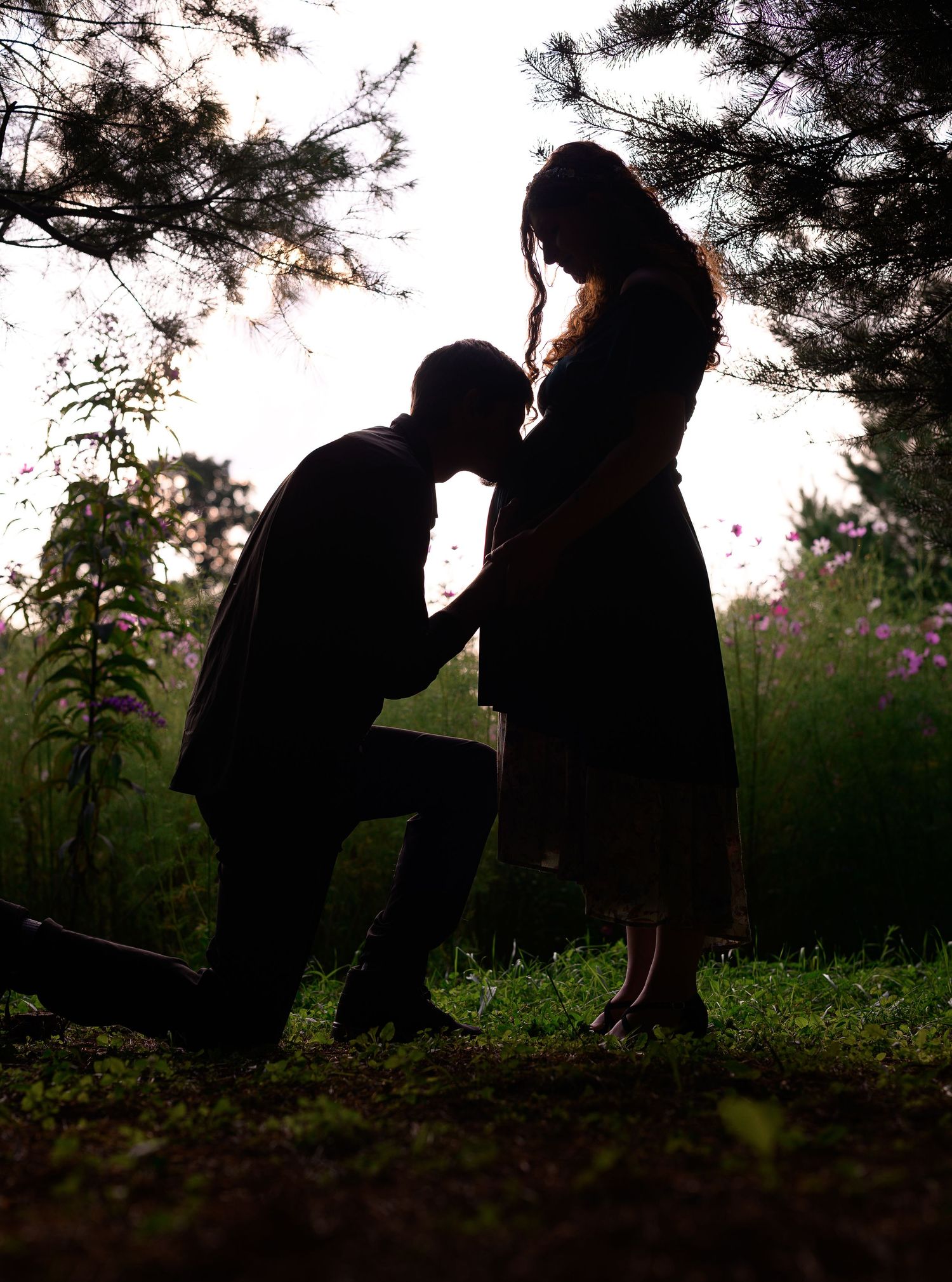 Silhouette of man kneeling to kiss pregnant woman's belly outdoors among trees and wildflowers at dusk.