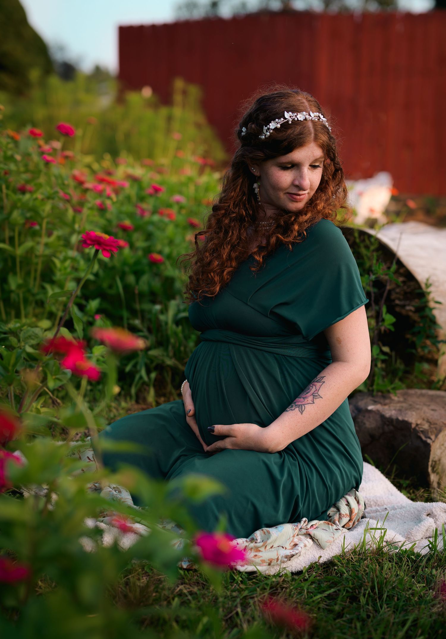 Pregnant woman in dark green dress sitting among pink flowers, wearing floral headband and cradling her baby bump.