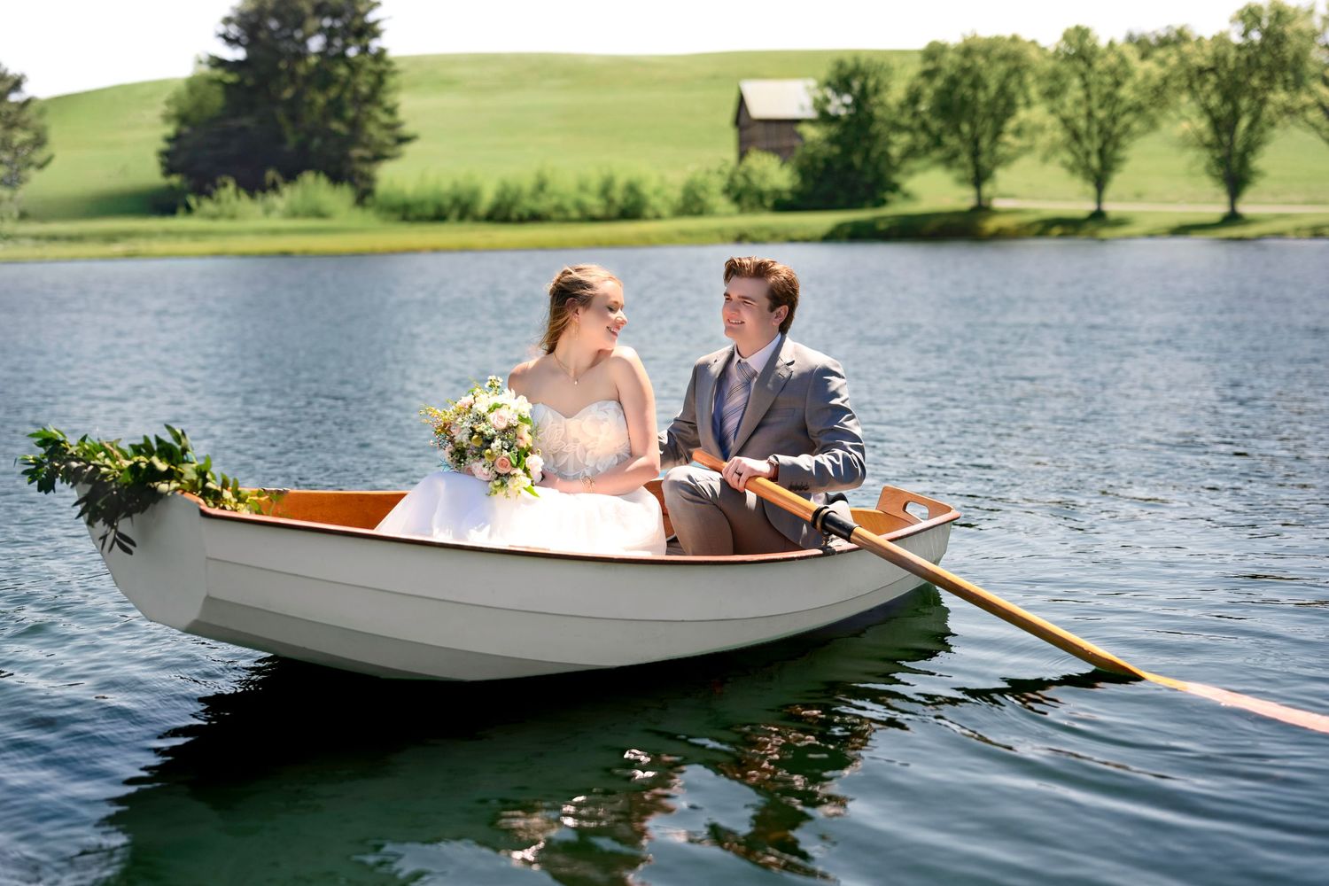 Bride and groom rowing on a serene lake in a white boat adorned with greenery, surrounded by lush scenery.