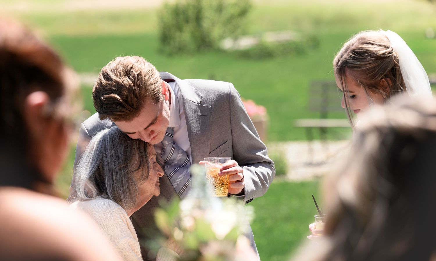 Bride and groom share a tender moment during outdoor wedding ceremony, groom in gray suit holding a glass.