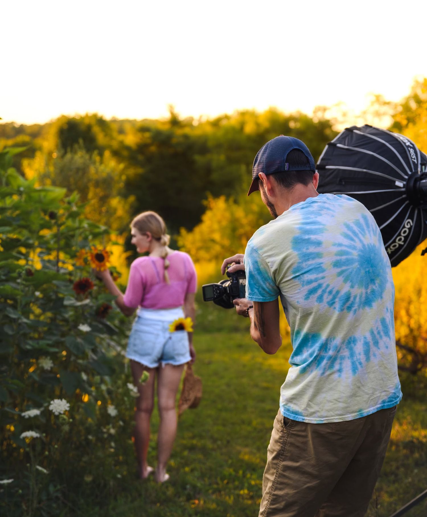 Photographer in tie-dye shirt captures woman picking sunflowers in golden hour field.