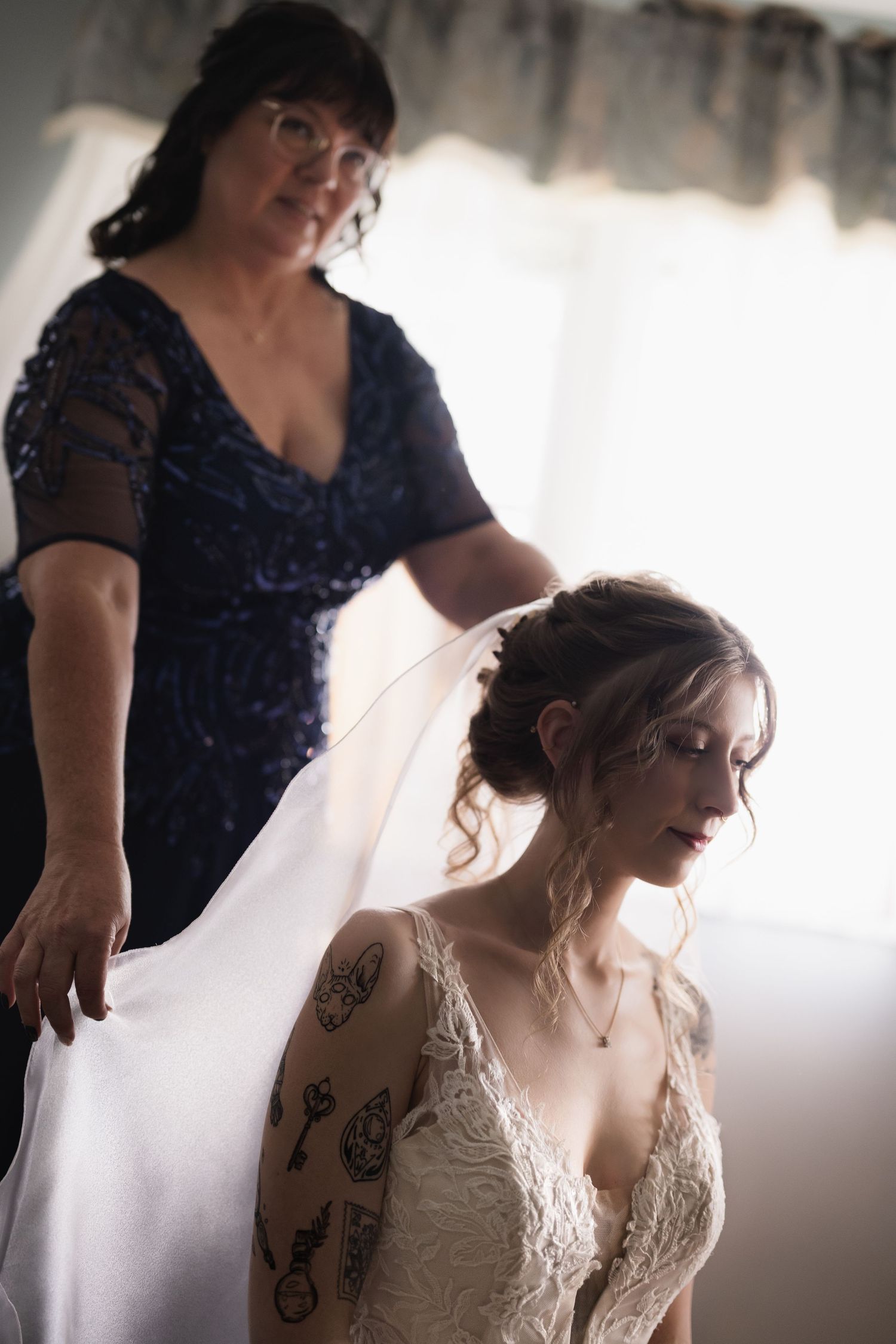 A bride with tattoos sits as an older woman in blue adjusts her white wedding veil near a bright window.
