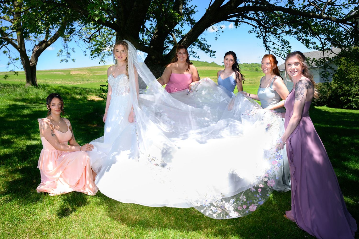 Bride in white gown with veil held by bridesmaids in colorful dresses outdoors under a tree on a sunny day.