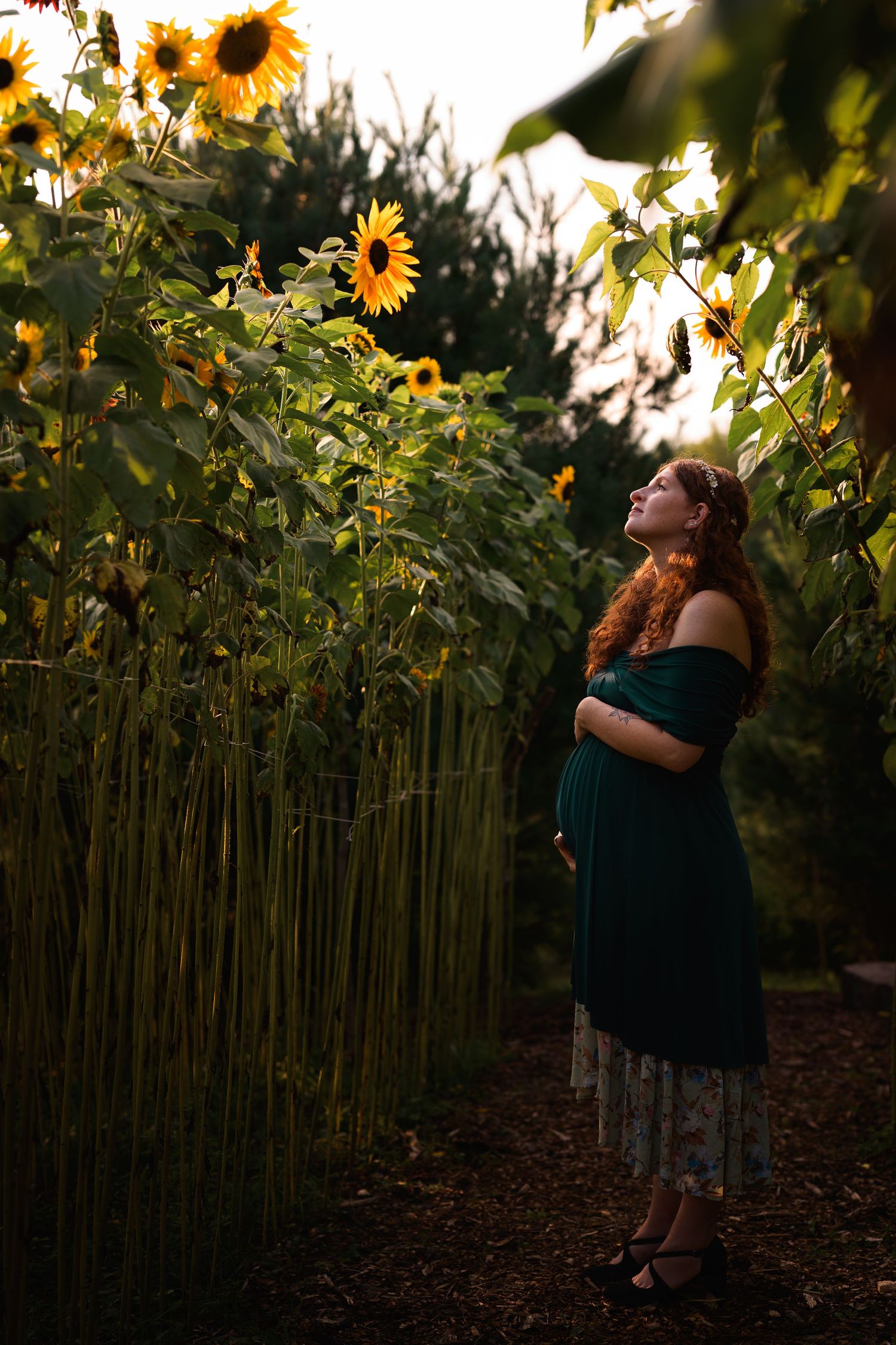 Pregnant woman in emerald off-shoulder dress standing among tall sunflowers, cradling baby bump in golden evening light.