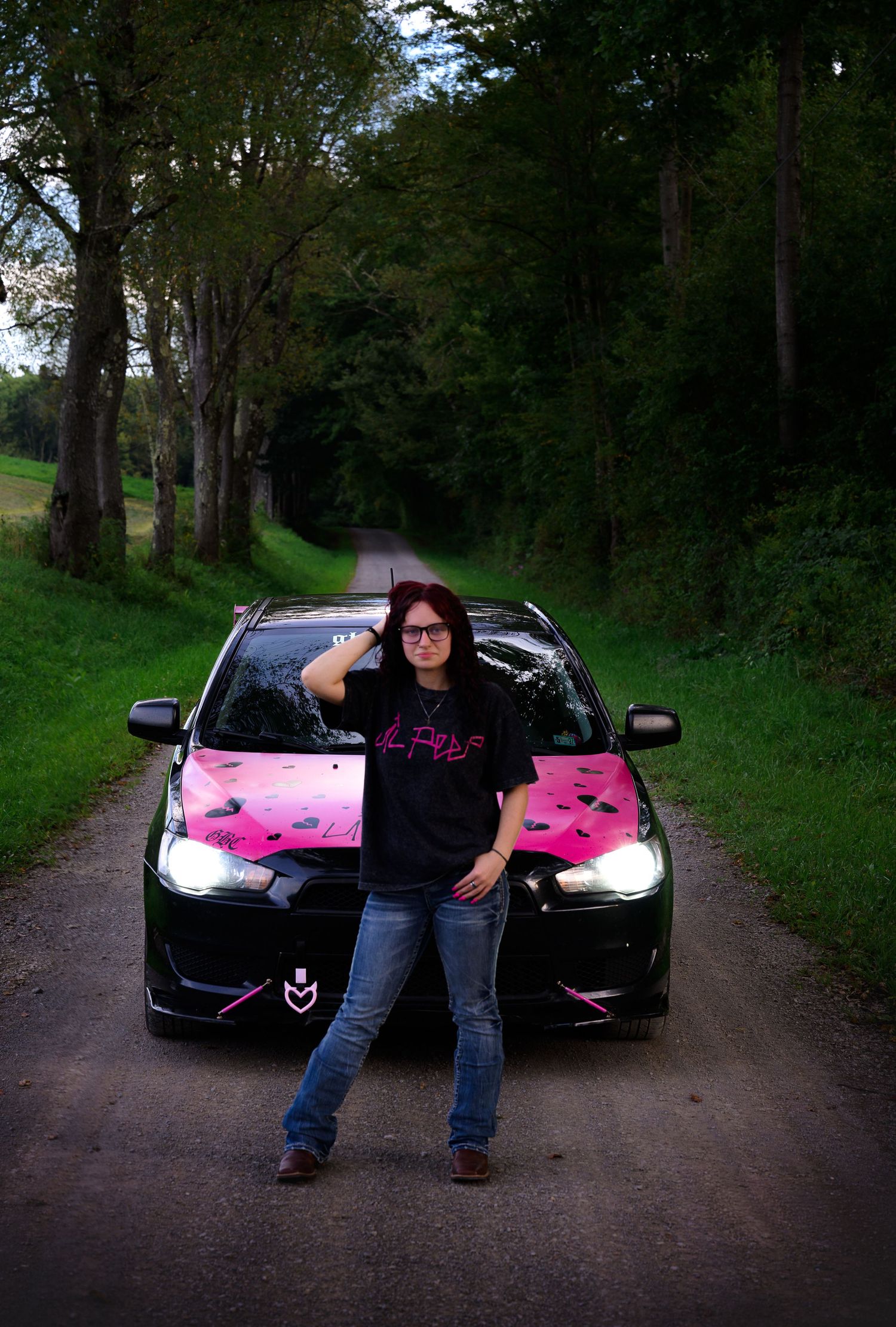 Woman in black graphic tee poses confidently in front of a black car with pink decals on a tree-lined road.