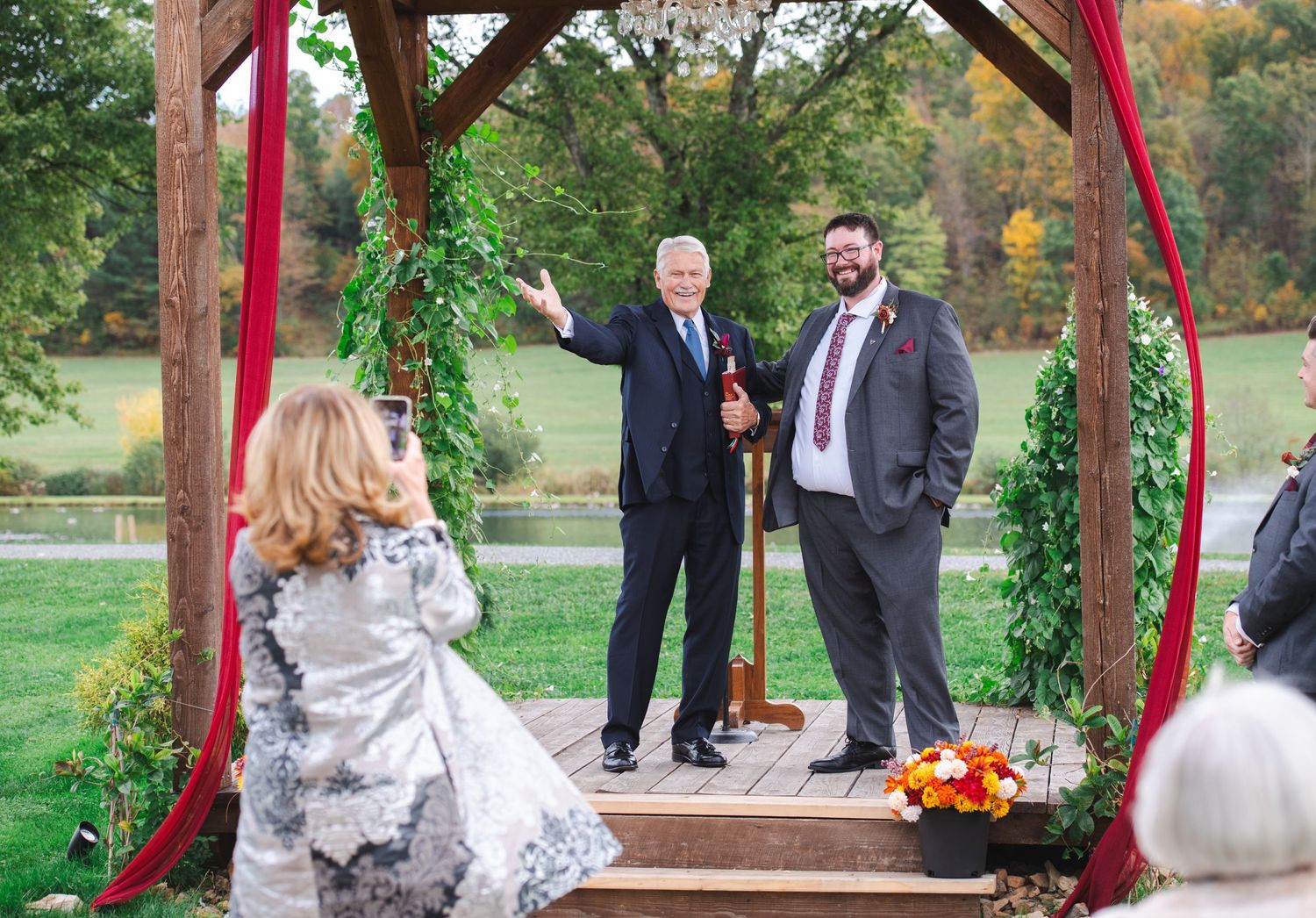 Officiant waves beside groom under rustic wooden wedding arch decorated with greenery and red ribbon outdoors.