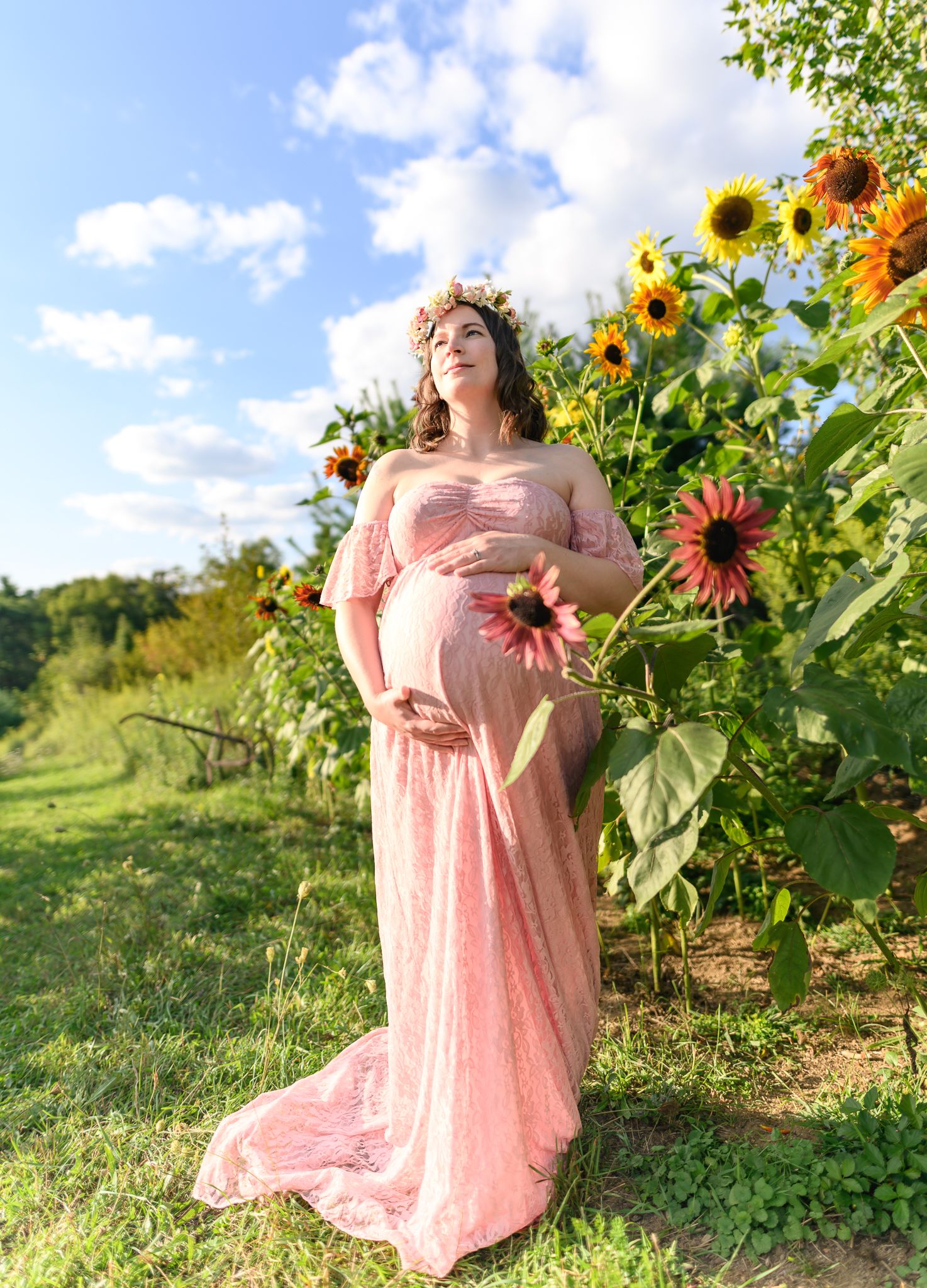 Pregnant woman in elegant pink lace maternity gown and floral crown posing among vibrant sunflowers outdoors.