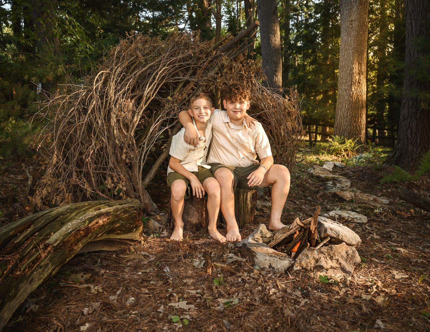 Two boys sit smiling inside a rustic twig fort in a sunlit forest setting.