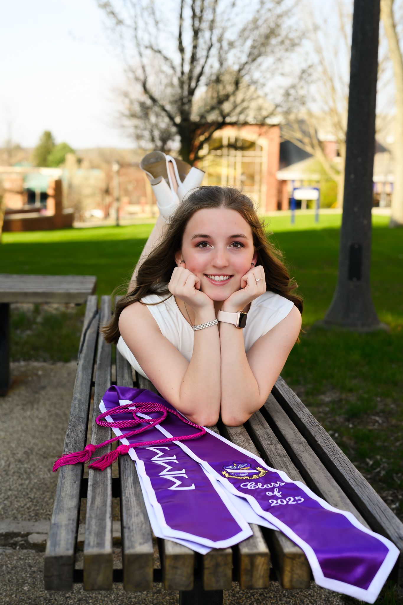 Smiling young woman wearing a crown and Class of 2023 sash poses on a park bench outdoors.