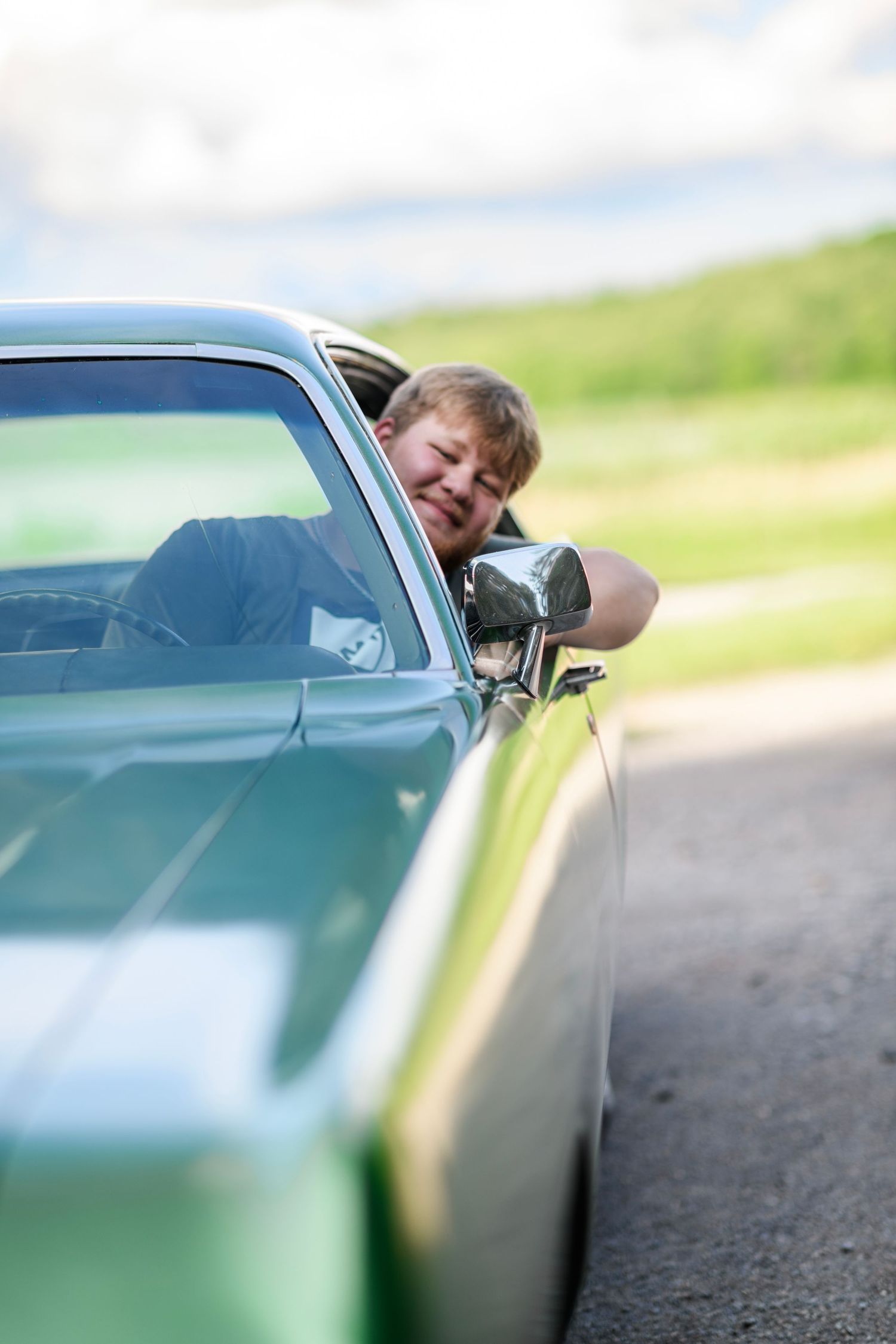 A smiling man leans out of a classic green car window on a sunny day with green fields in the background.