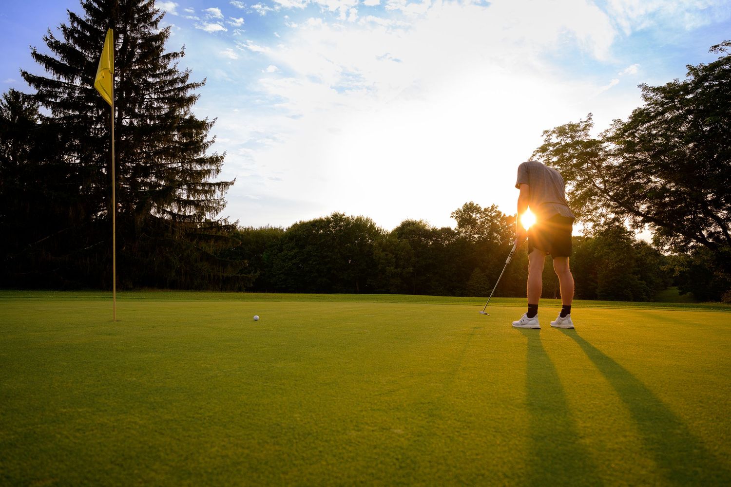 Golfer putting on a lush green golf course at sunset with a yellow flag and trees in the background.