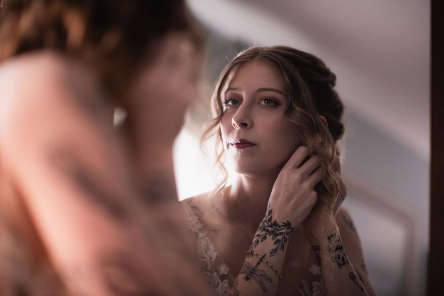 Bride adjusting earring while gazing into mirror, wearing floral lace gown in soft romantic lighting.