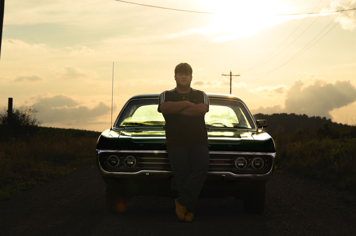 Man leaning against a classic black car at sunset on a rural road, arms crossed, golden sky in background.