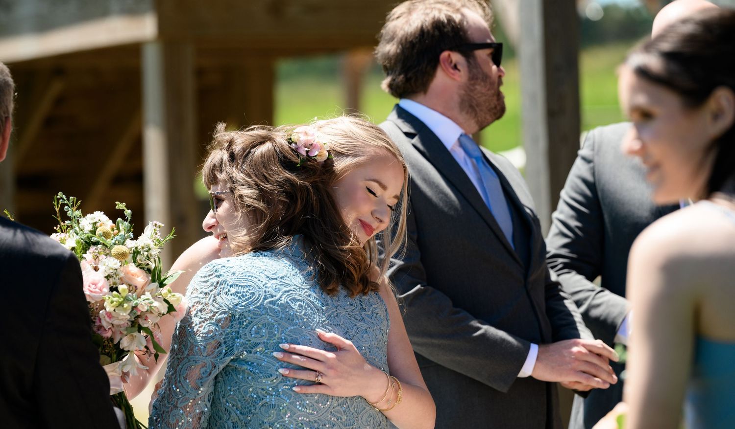 Two women share an emotional hug at an outdoor wedding ceremony, with guests standing nearby in formal attire.