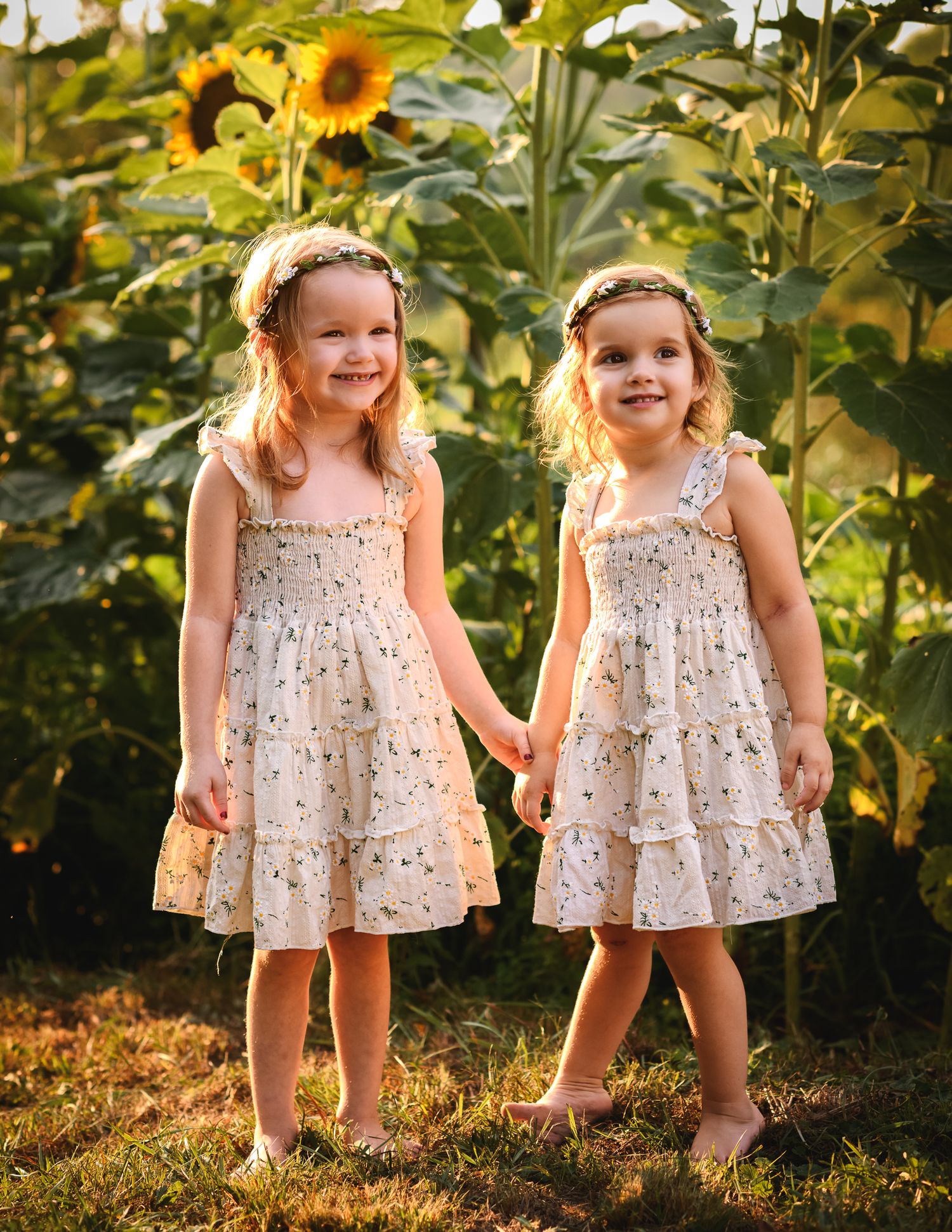 Two smiling twin girls in floral dresses hold hands in a sunflower field during golden hour.