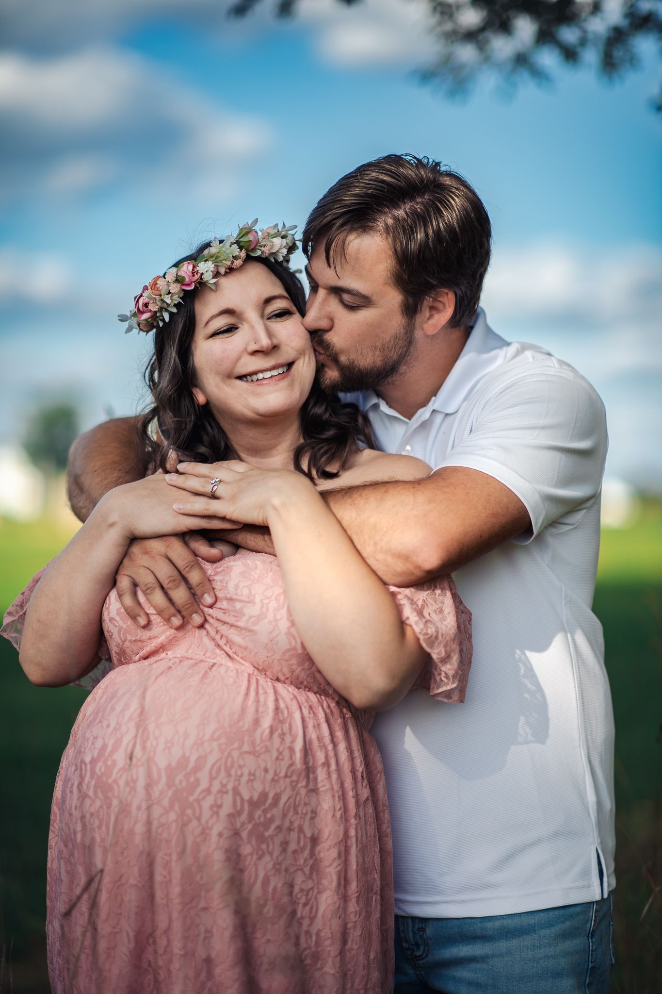 Pregnant woman in pink lace dress and floral crown embraced by man in white shirt outdoors.