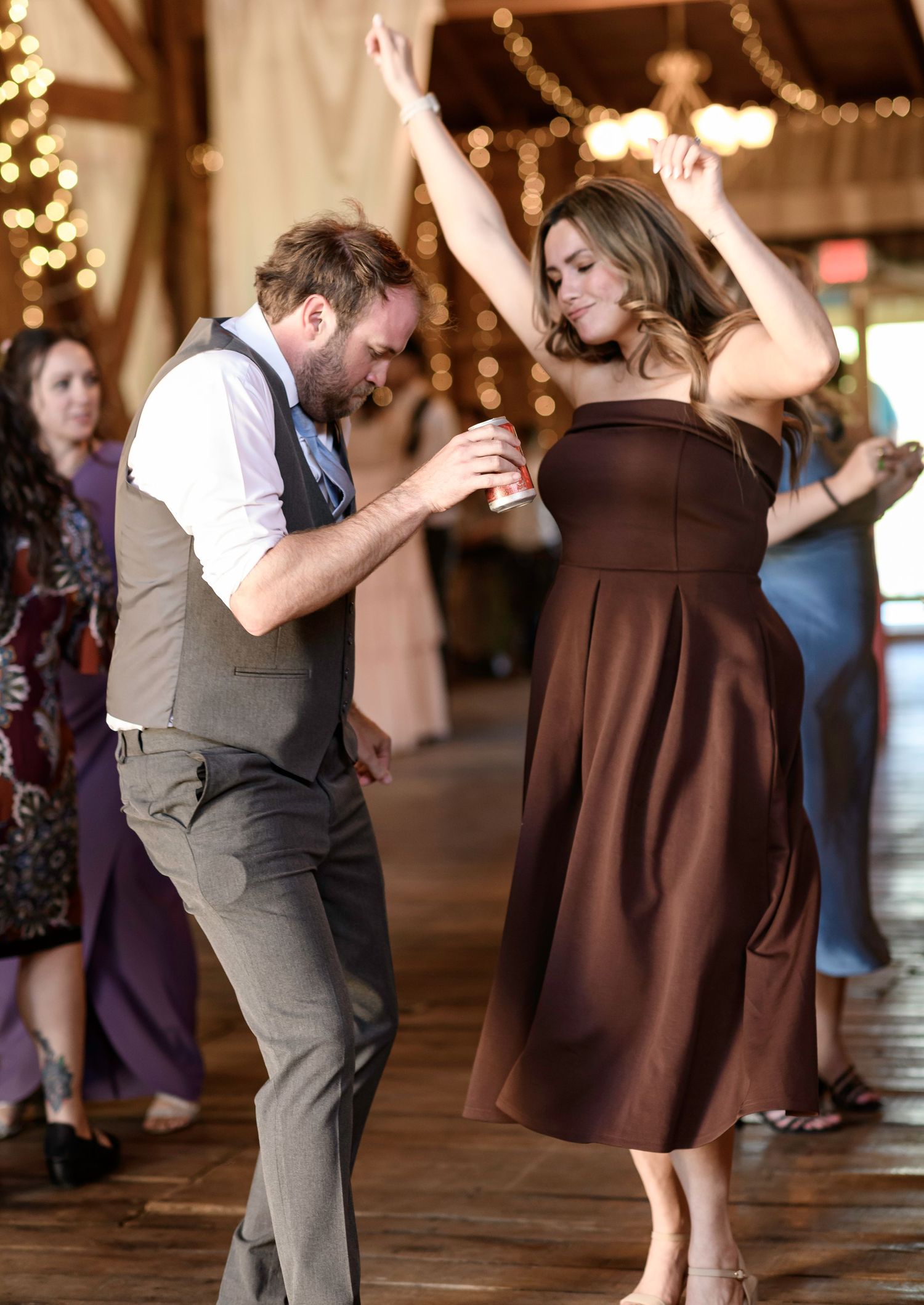 Couple dancing energetically at a wedding reception in a rustic barn venue with warm string lights.