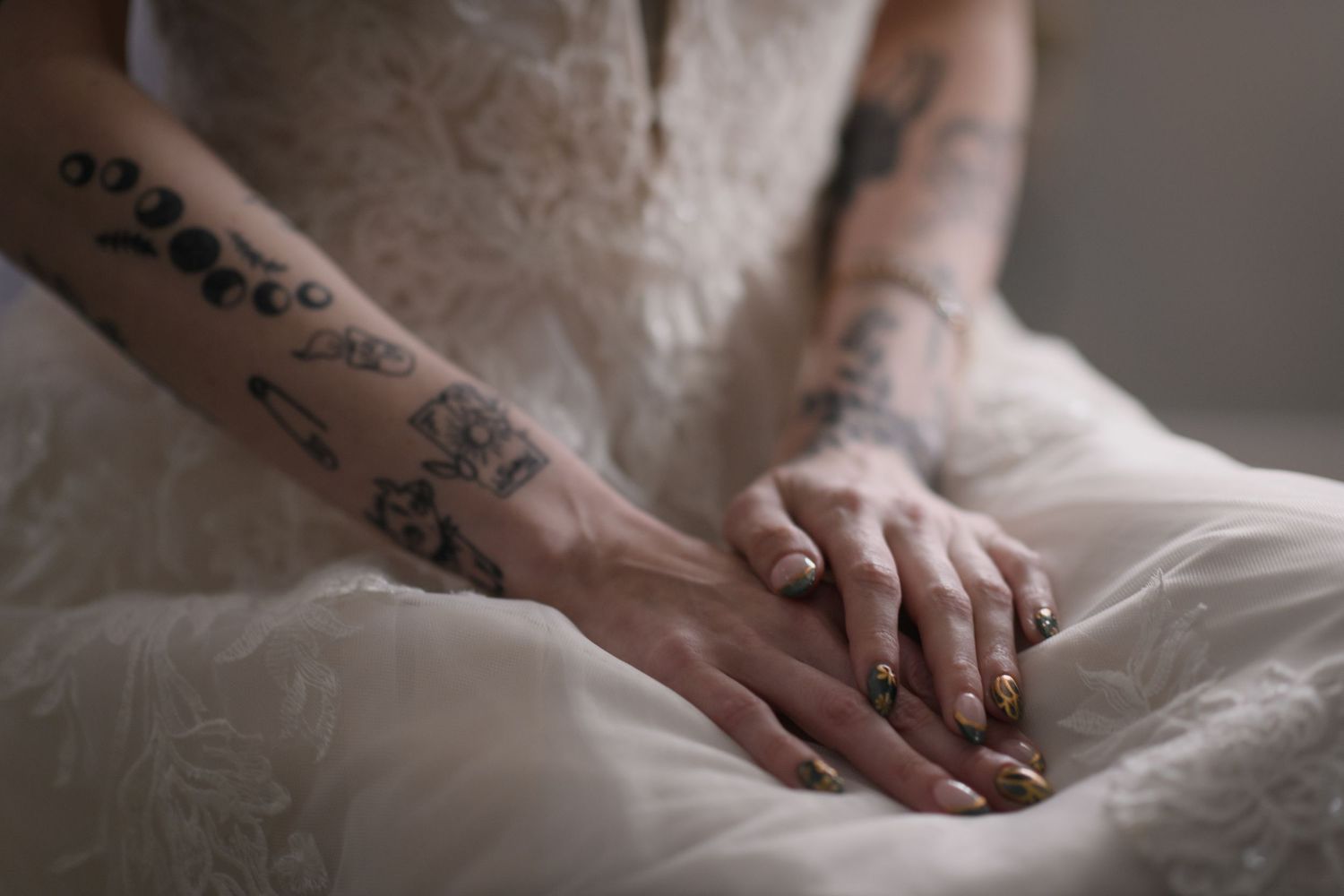 Tattooed bride resting hands on white wedding dress, wearing gold nail polish and wedding ring.