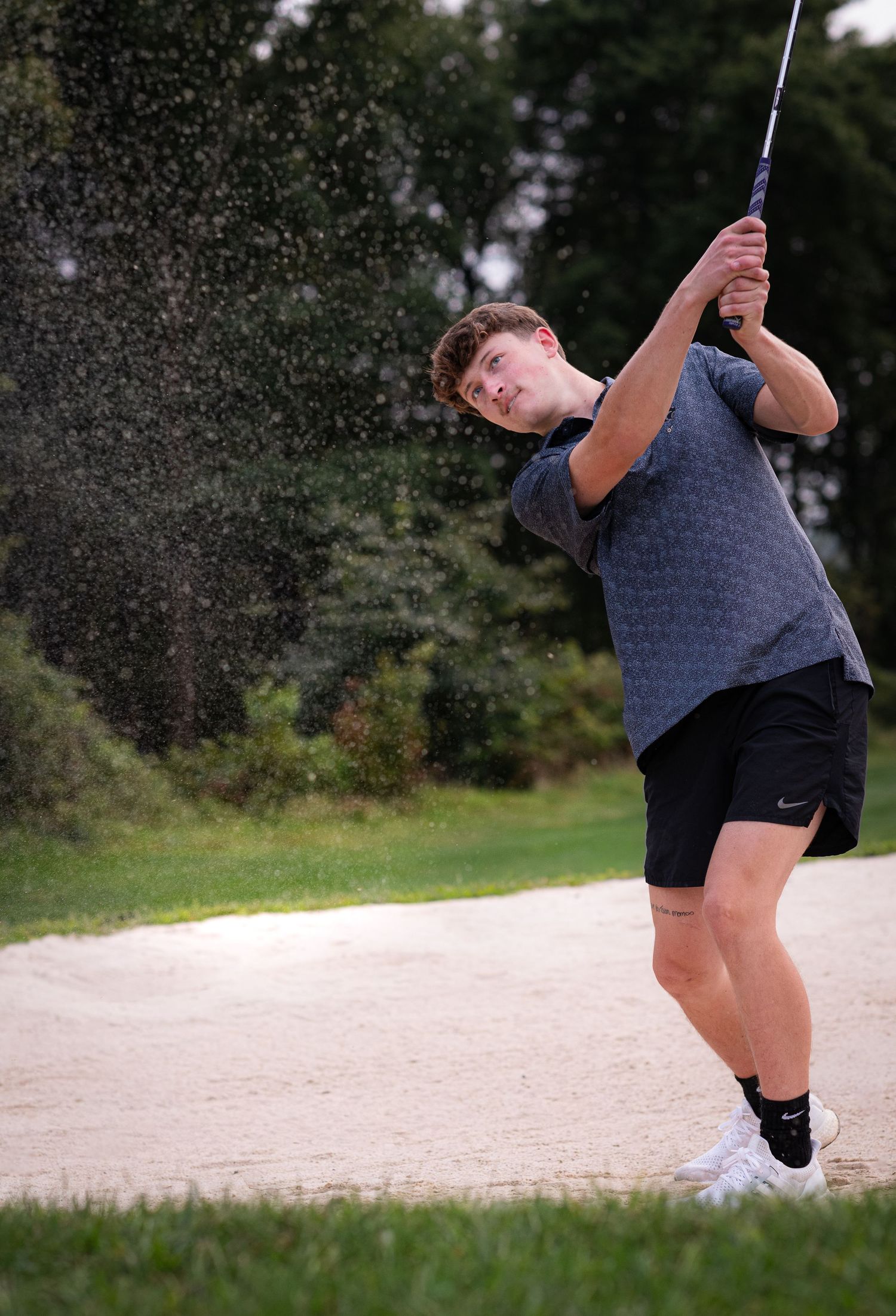 Young golfer swinging a club from a sand bunker on a golf course, sending sand flying through the air.
