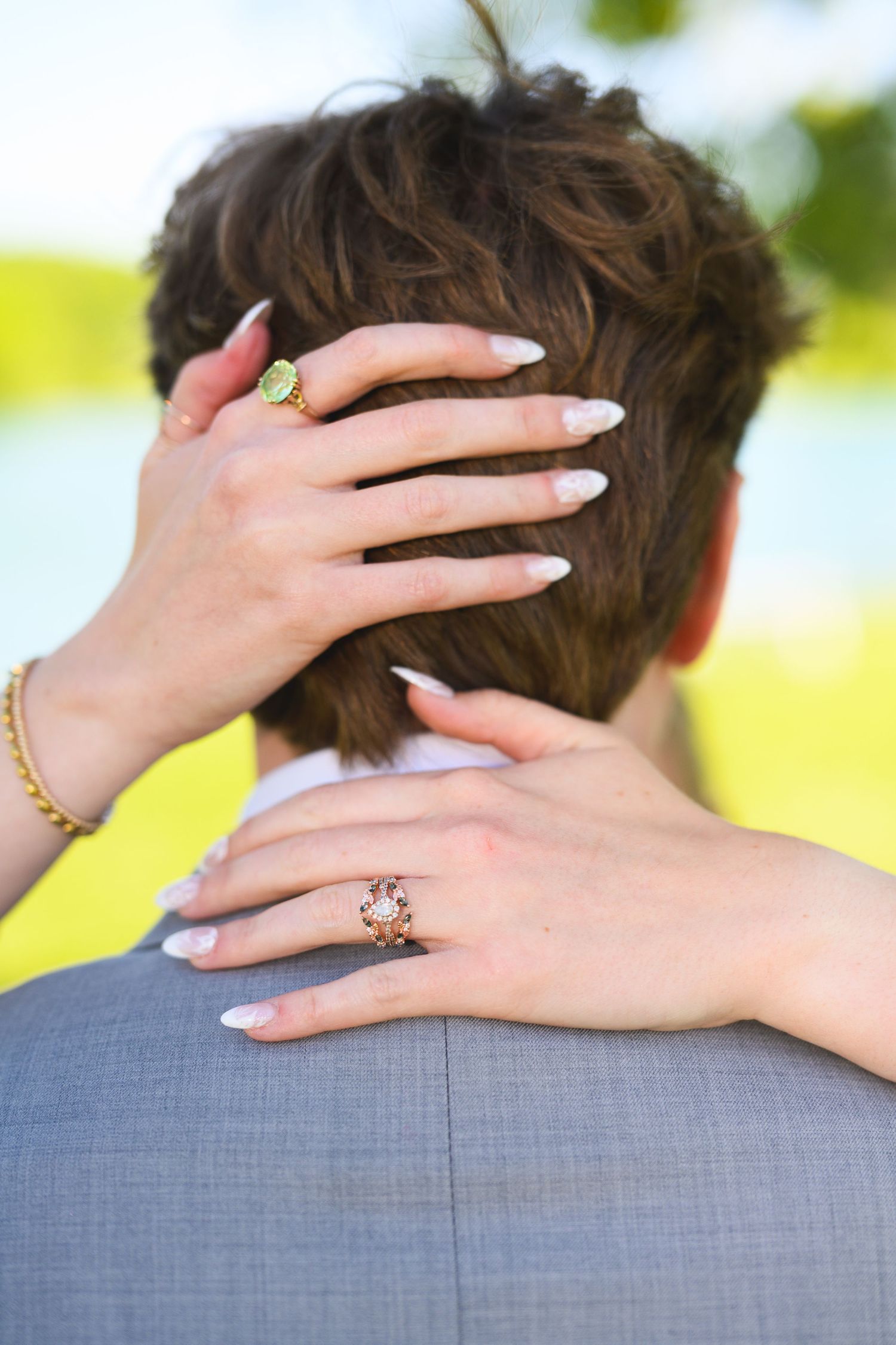 Woman with manicured nails and elegant rings embraces man in gray suit from behind outdoors.
