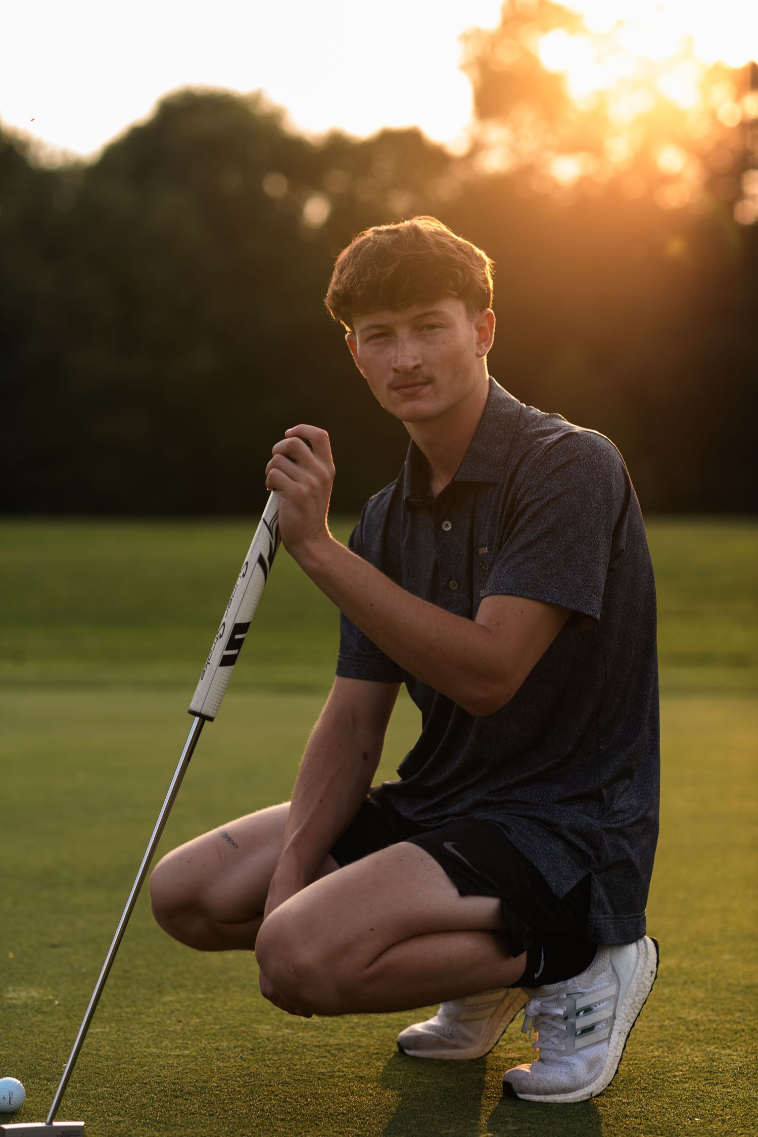 Young golfer kneeling on green golf course at golden sunset, holding putter club in dark polo shirt and shorts.