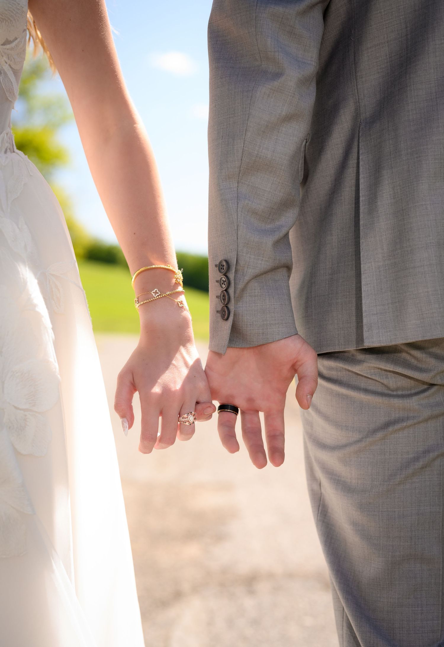 Bride and groom holding hands, showing wedding rings and gold bracelets, dressed in white gown and gray suit.