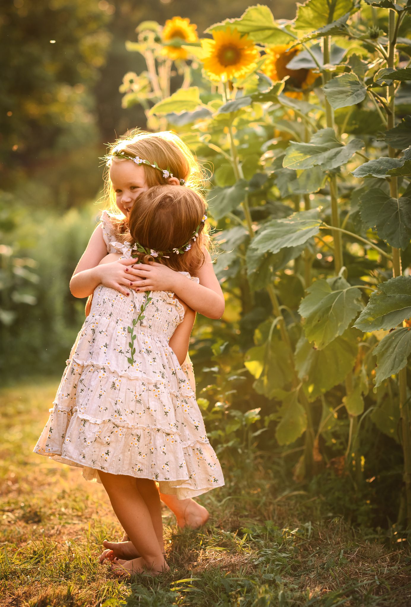 Two young girls in floral dresses and flower crowns share a sweet hug among tall sunflowers in golden light.