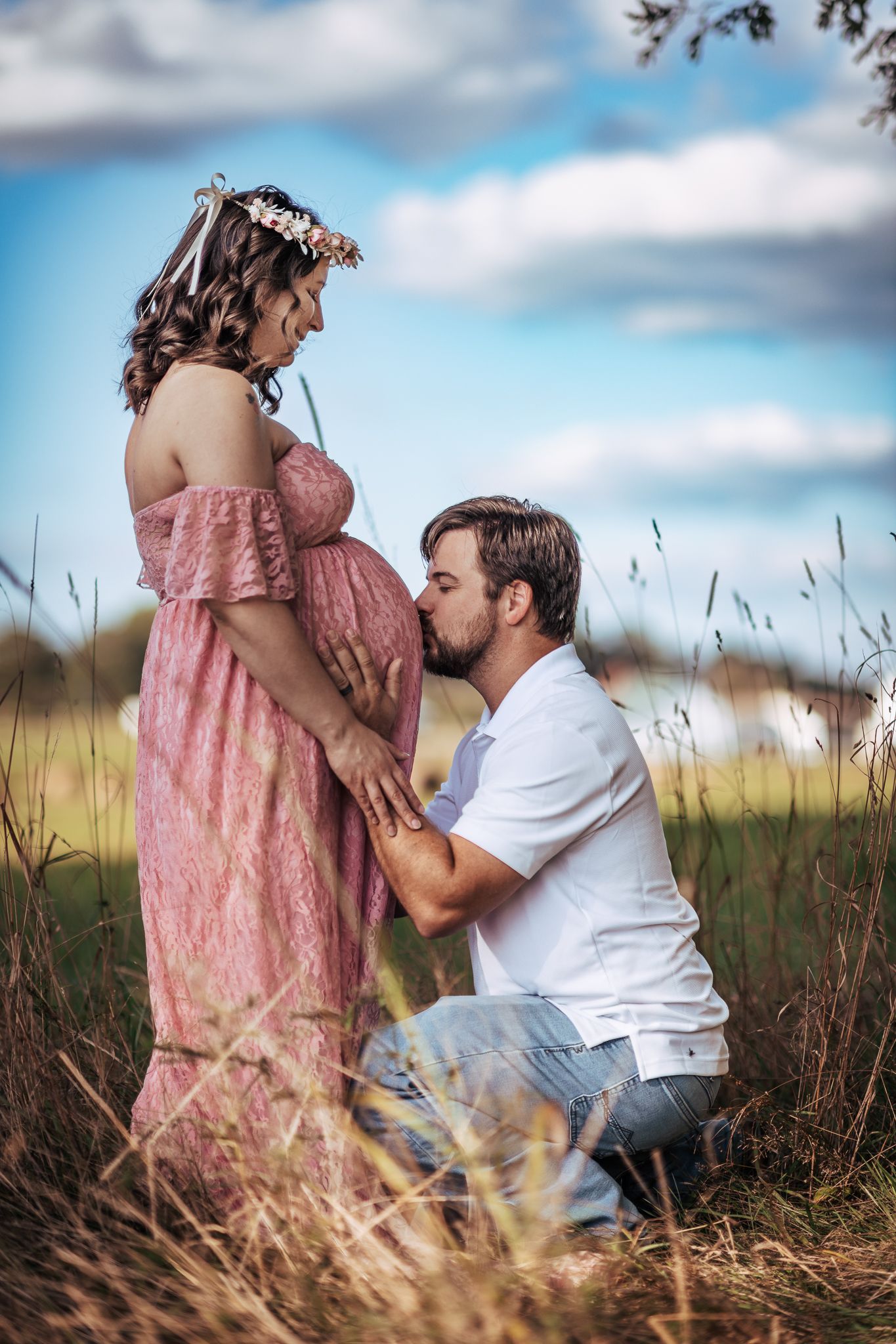 A man kneels in a field kissing a pregnant woman's belly, as she wears a pink dress and floral crown.