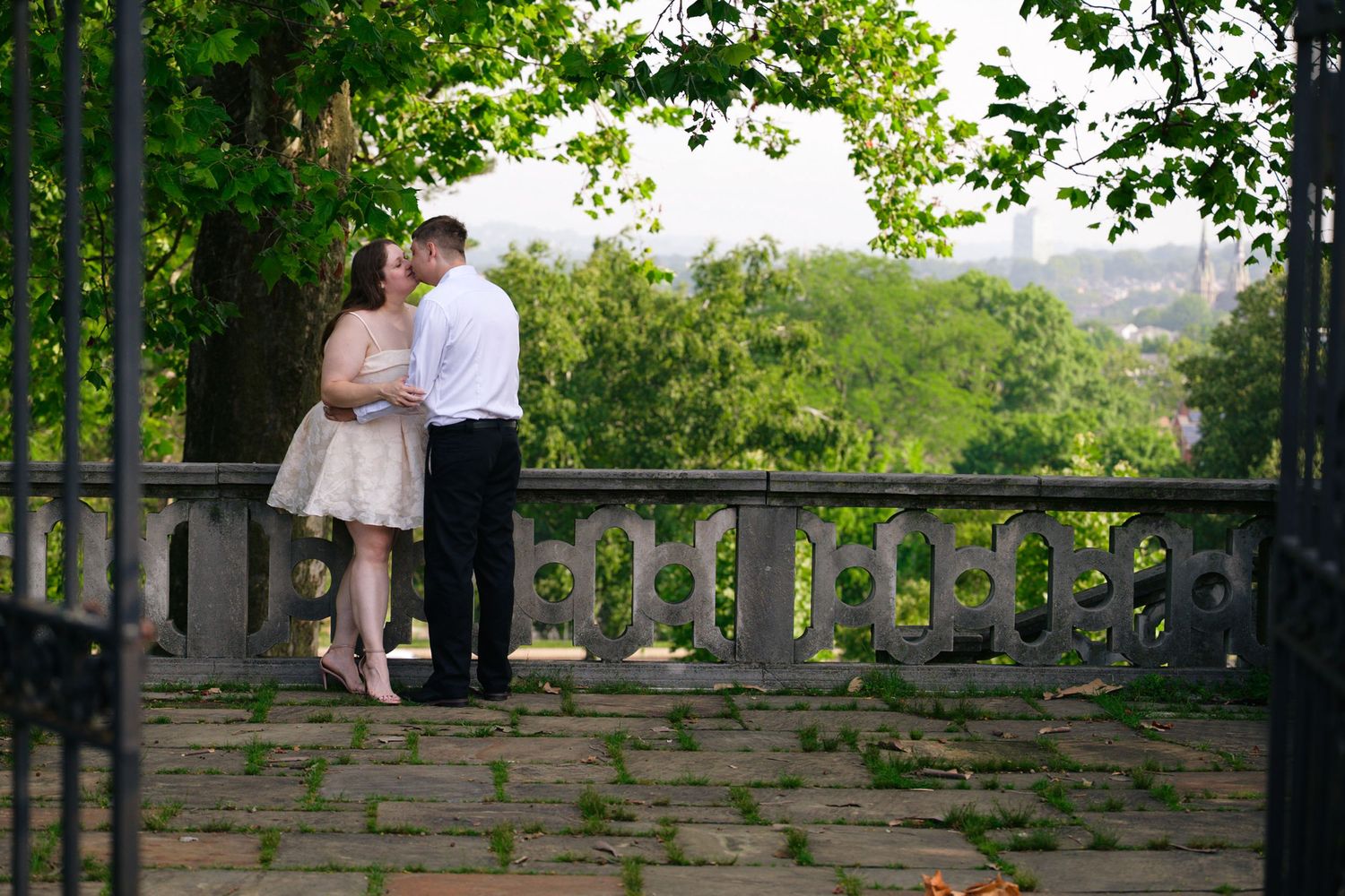 A couple shares a romantic kiss on a stone terrace surrounded by lush green trees in an outdoor garden setting.