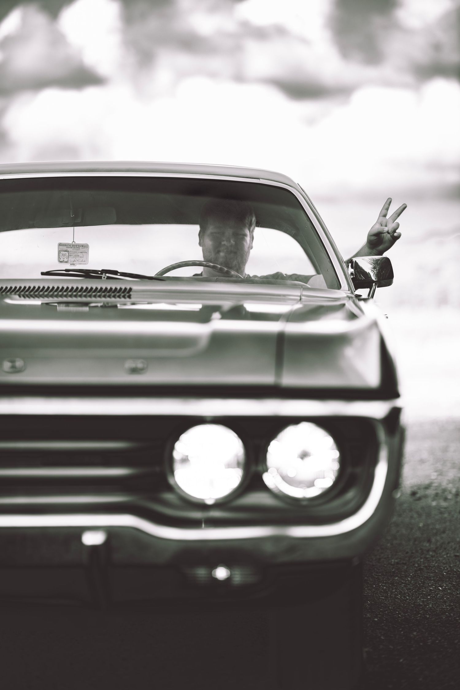 Black and white photo of a person flashing a peace sign from inside a classic vintage muscle car with round headlights.