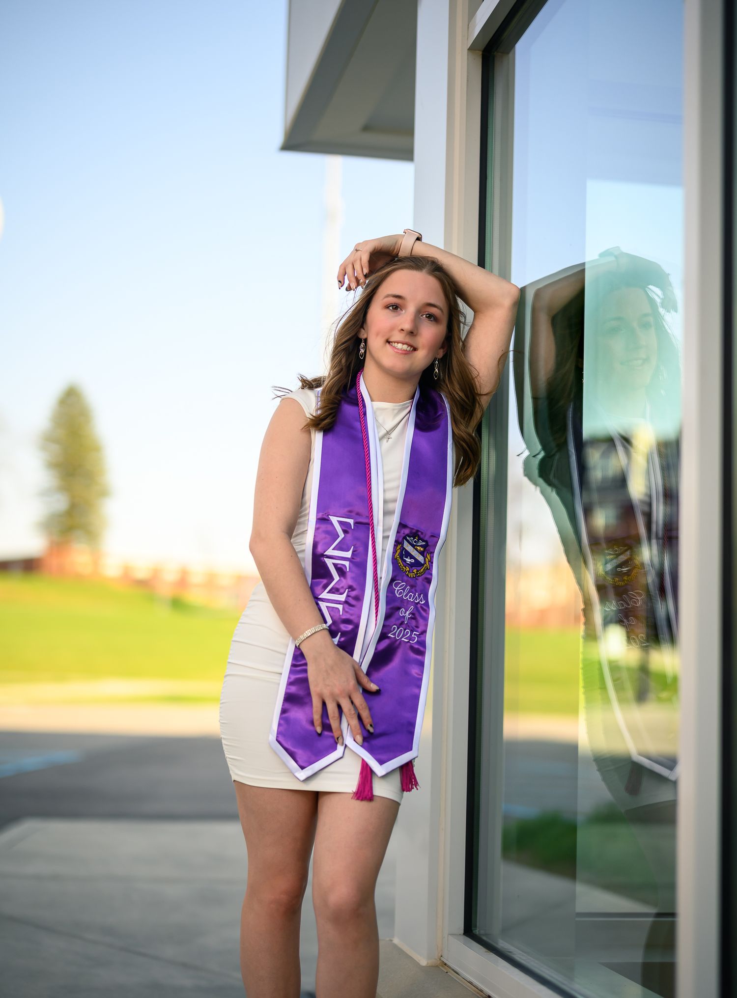 Young woman in white dress and purple graduation stole poses confidently outdoors during golden hour senior photo session.