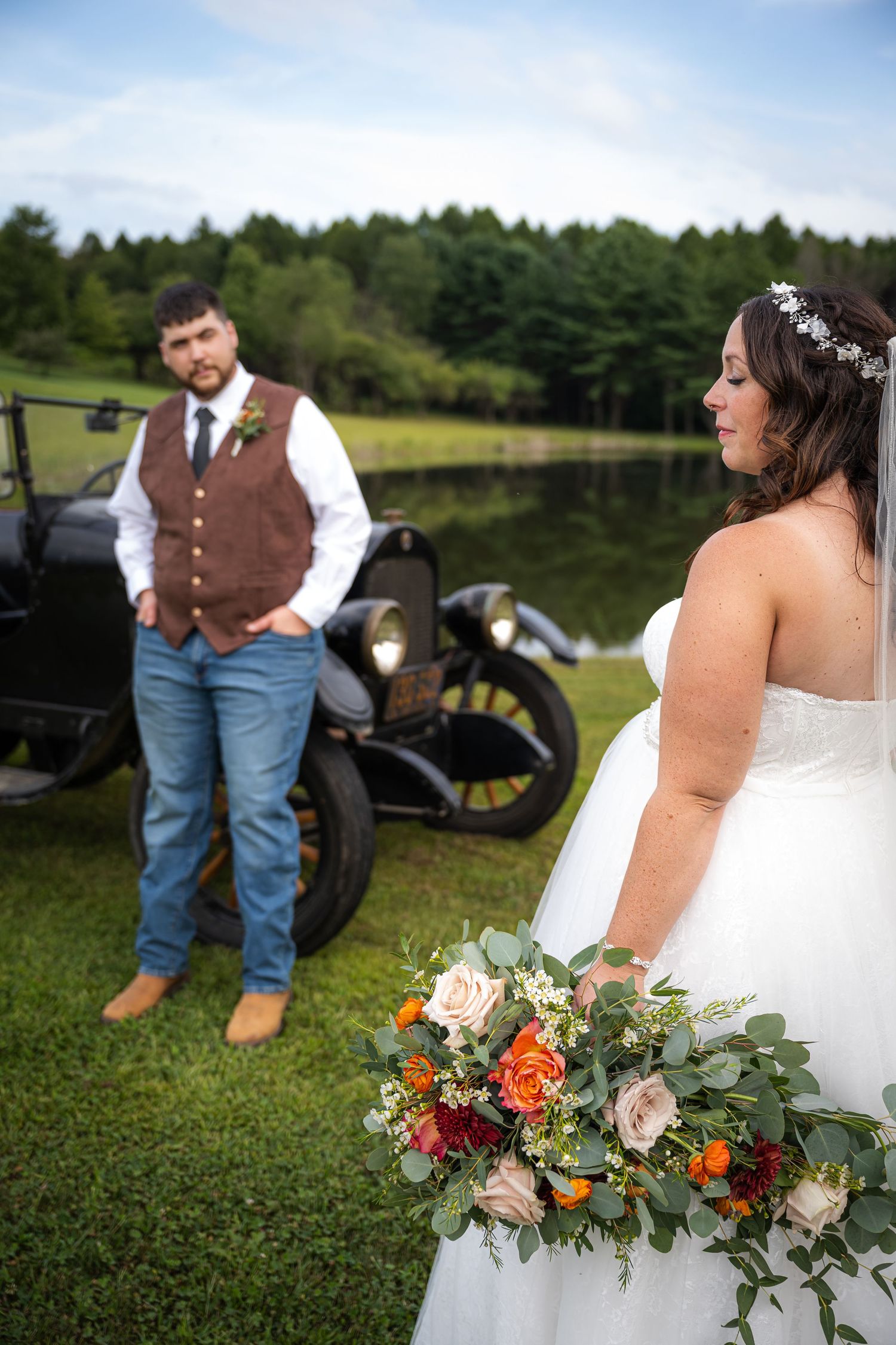 Bride holding colorful bouquet gazes at groom leaning on vintage black car at outdoor wedding.
