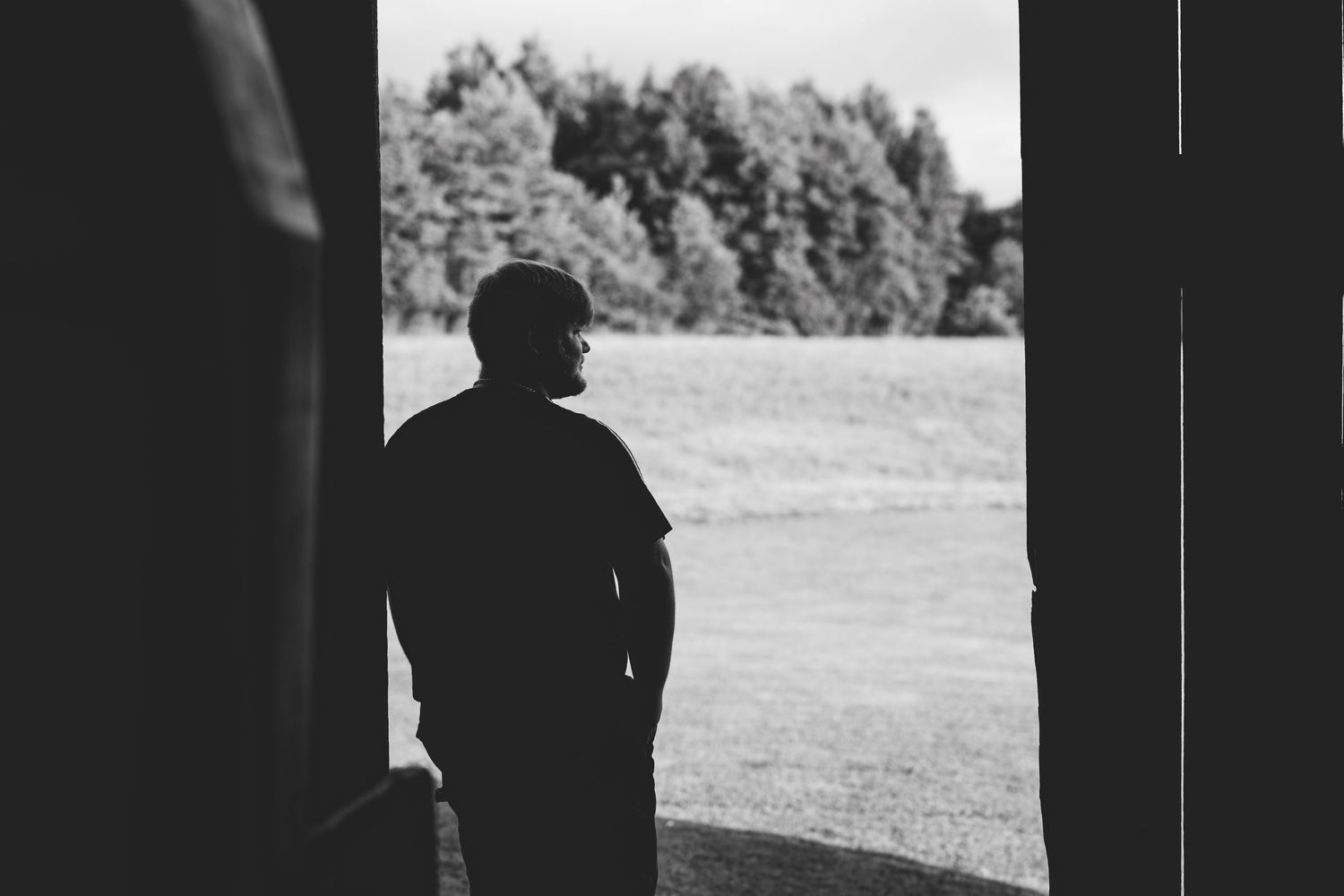 Black and white photo of a man standing in a doorway gazing out at a lush green field surrounded by trees.