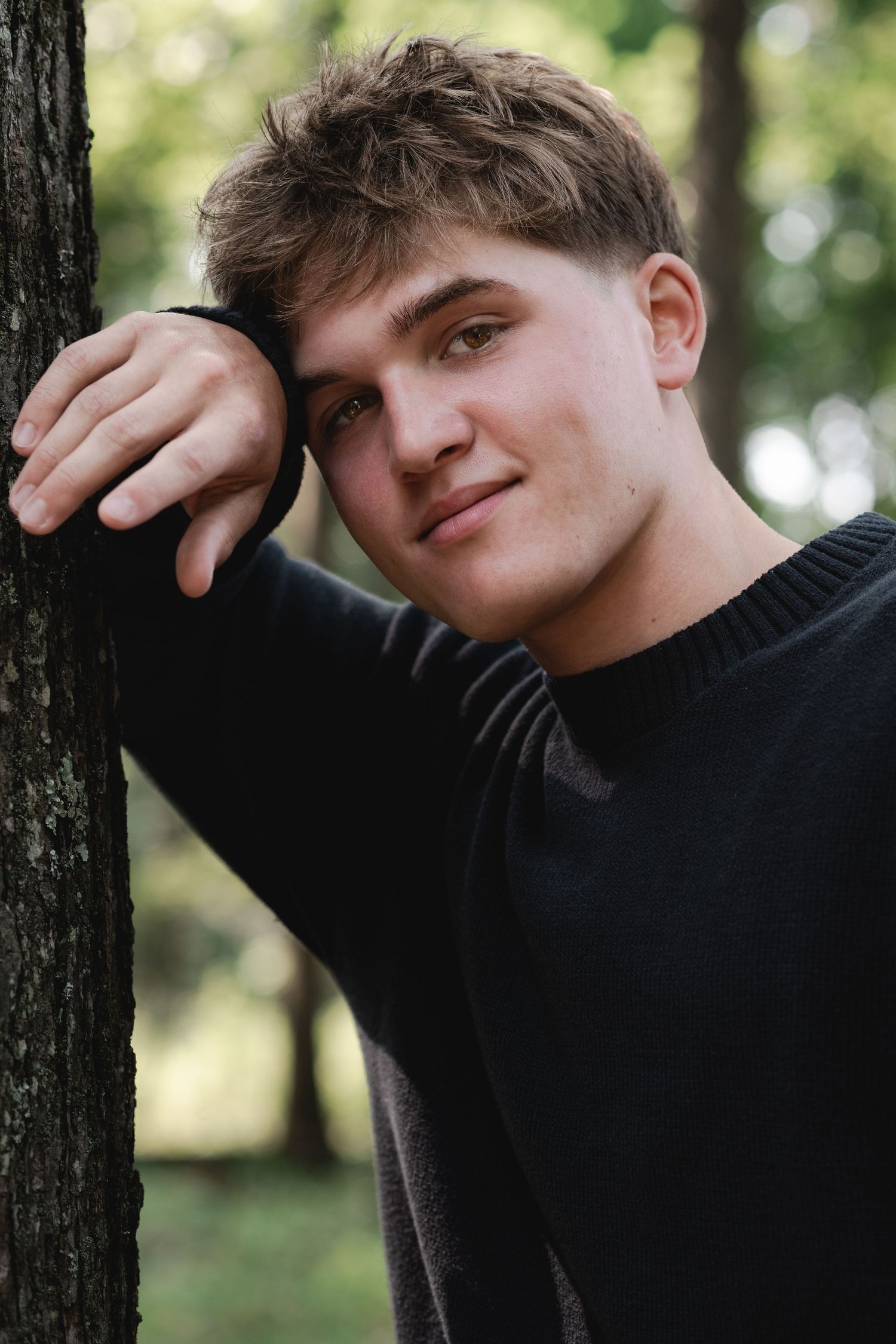 Young man in black sweater leaning against a tree in a park, smiling softly at the camera.
