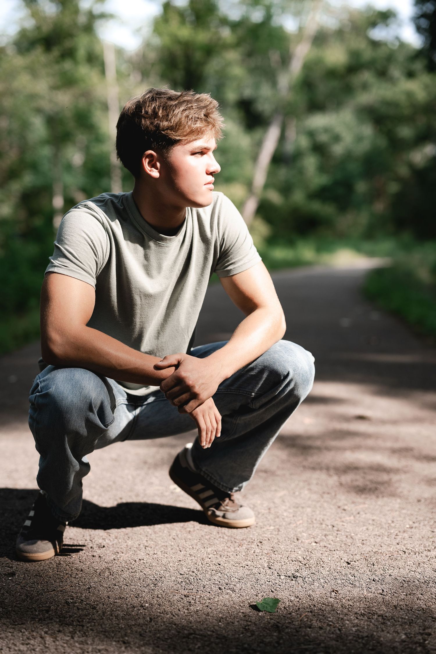 Young man in a gray t-shirt and jeans crouching on a path surrounded by green trees.