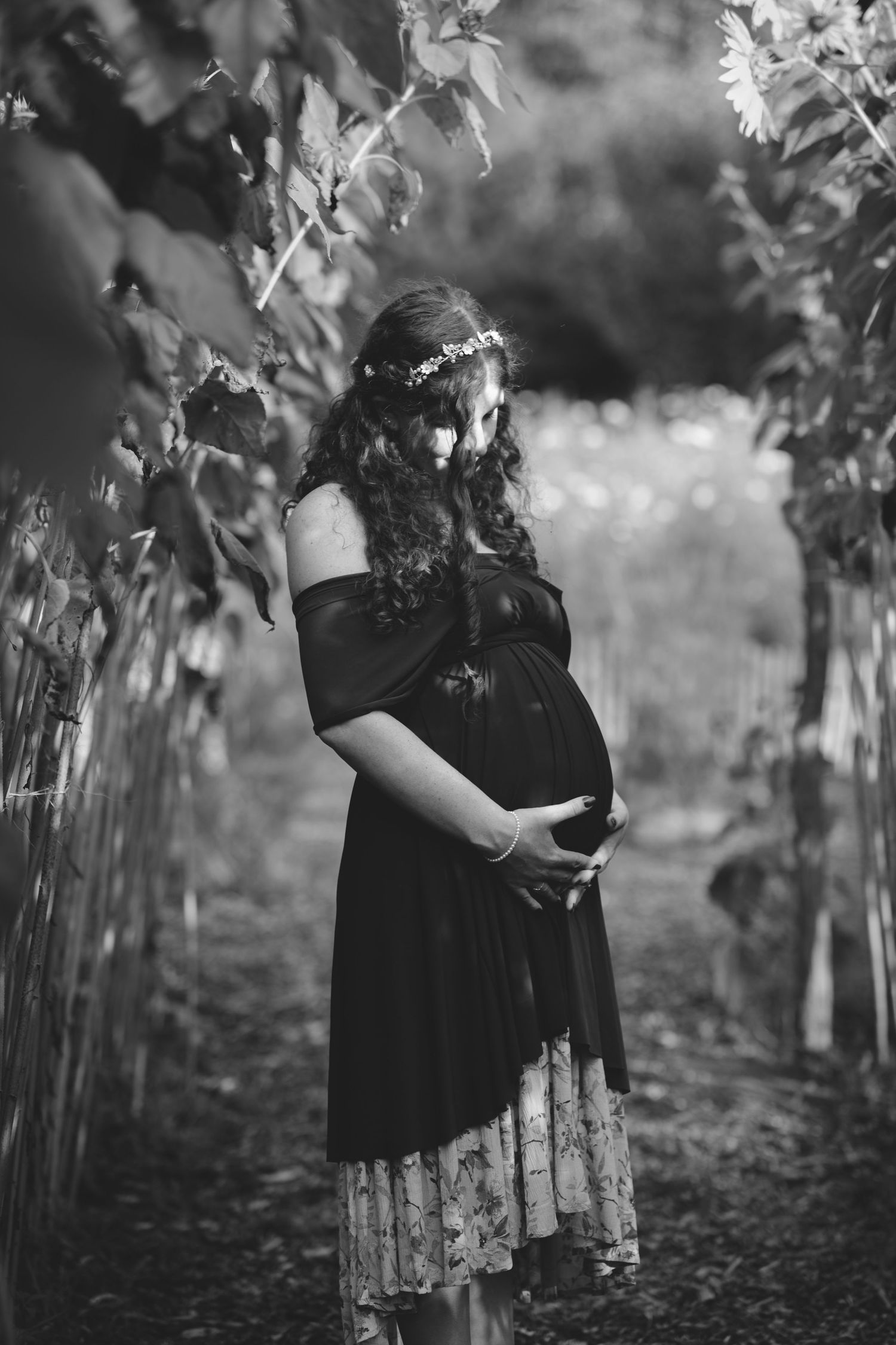 Black and white photo of a pregnant woman in a floral crown holding her belly in a lush garden pathway.