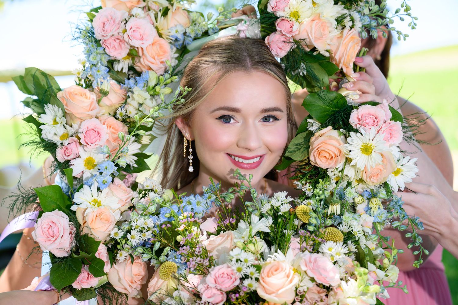Young blonde woman smiling through a wreath of pink roses, daisies, and blue hydrangeas at an outdoor event.