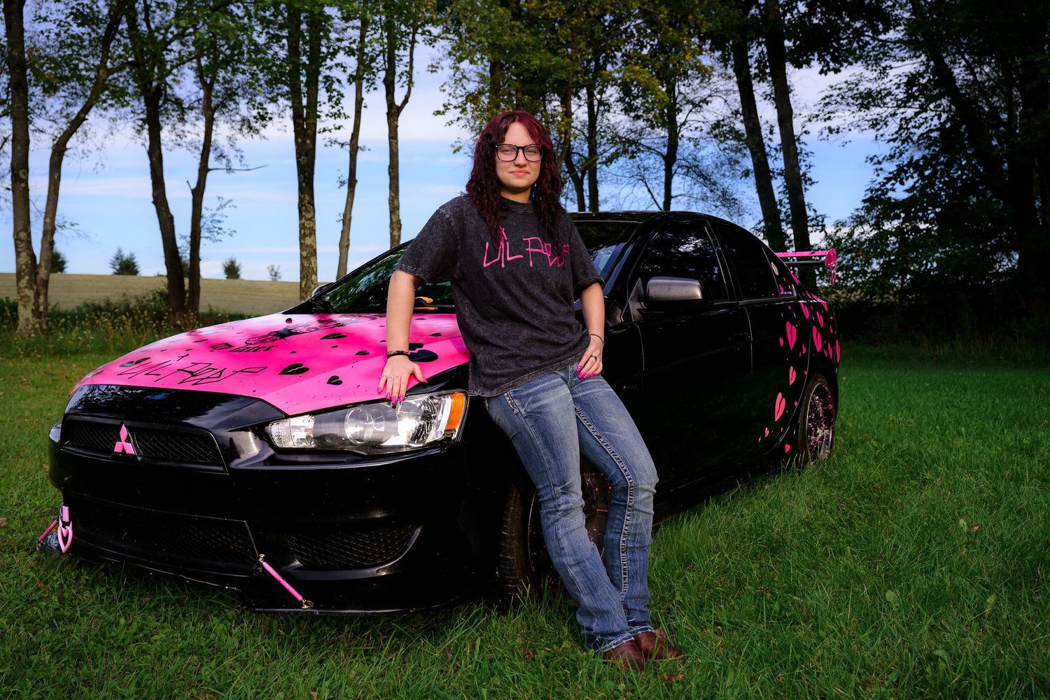 Woman leaning on black and pink Mitsubishi car with breast cancer awareness ribbons outdoors.