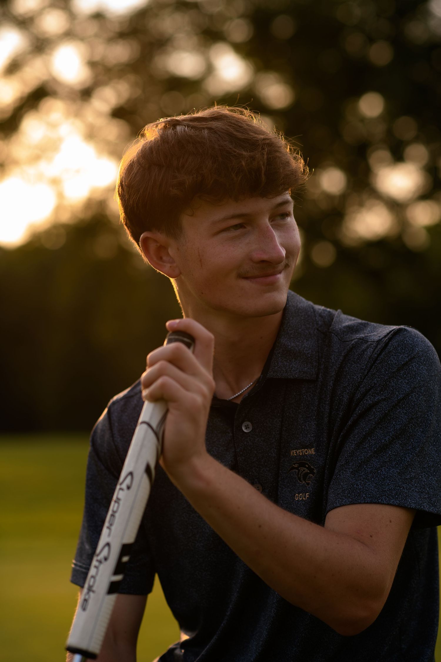 Young golfer holding a club at golden hour, smiling outdoors on a golf course at sunset.