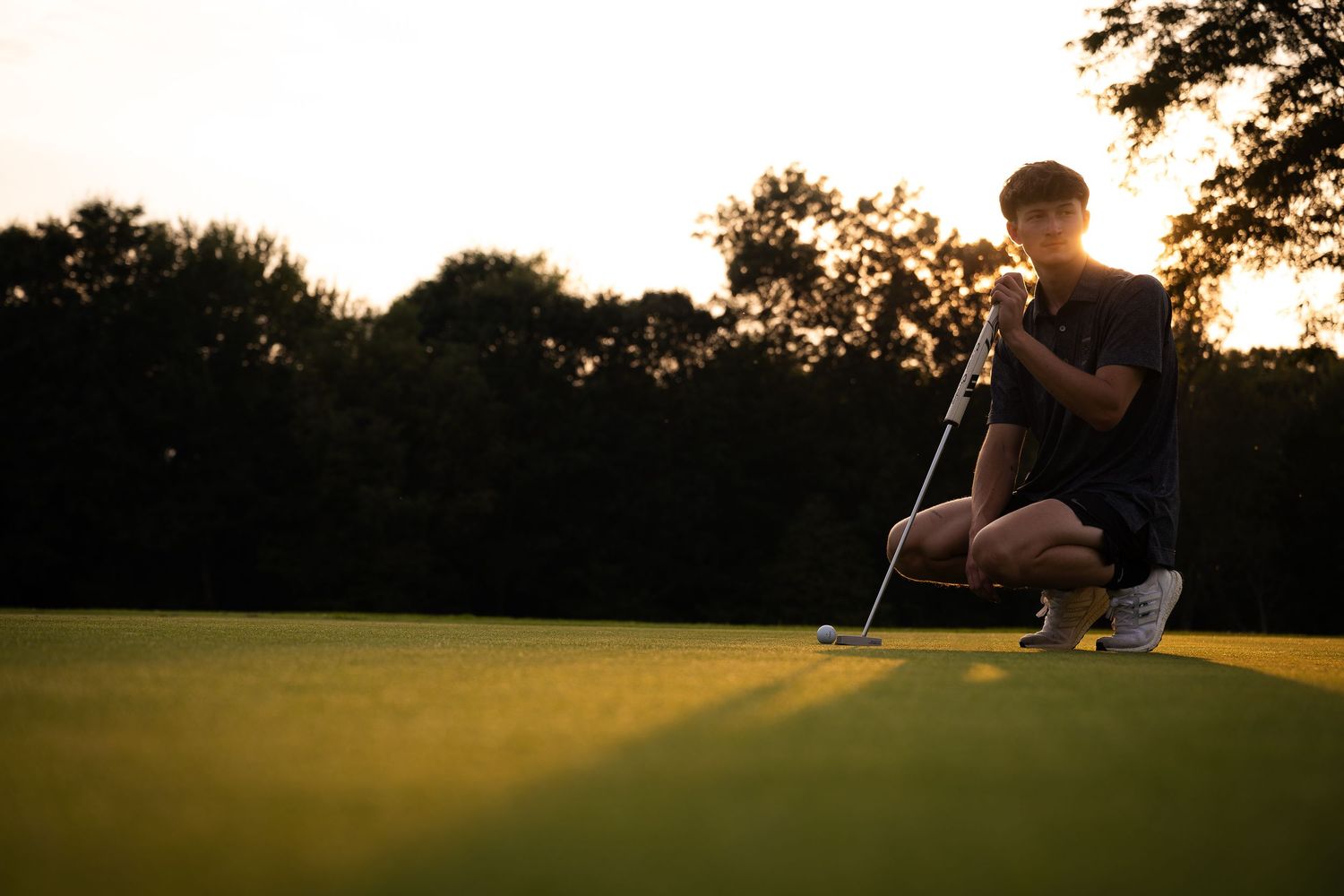 Golfer crouching on a lush green putting green at sunset, analyzing the line of a putt with golf club in hand.