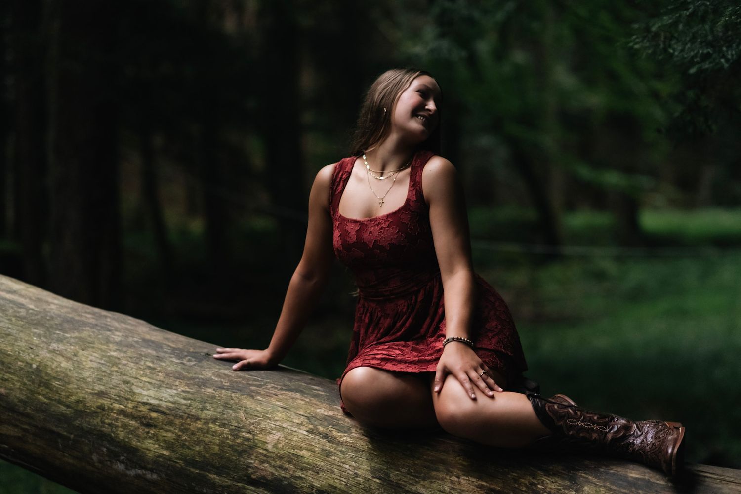 Woman in red lace dress and cowboy boots sits on a fallen log in a lush green forest, smiling.