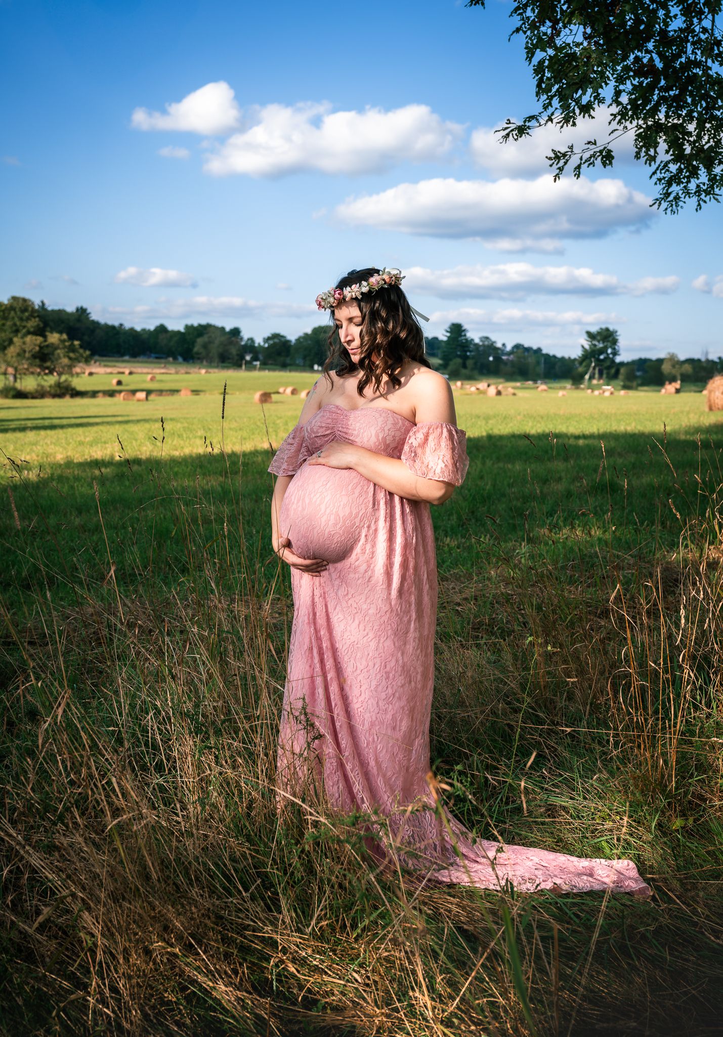 Expectant mother in pink lace maternity dress with floral crown poses in sunny green meadow.