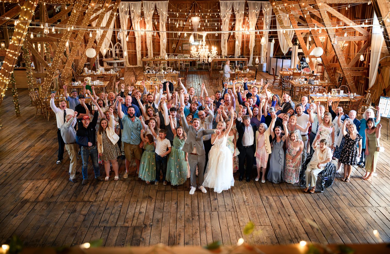 Joyful wedding party group photo with all guests raising hands in a beautifully decorated rustic barn.