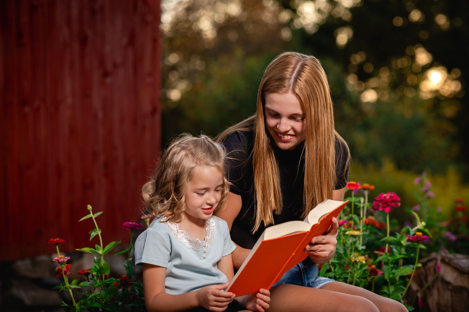 Older girl and young child reading an orange book together outdoors near red barn and flowers.