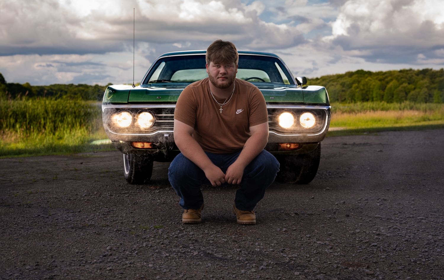 Young man crouching in front of a classic green muscle car on a rural road under dramatic cloudy skies.
